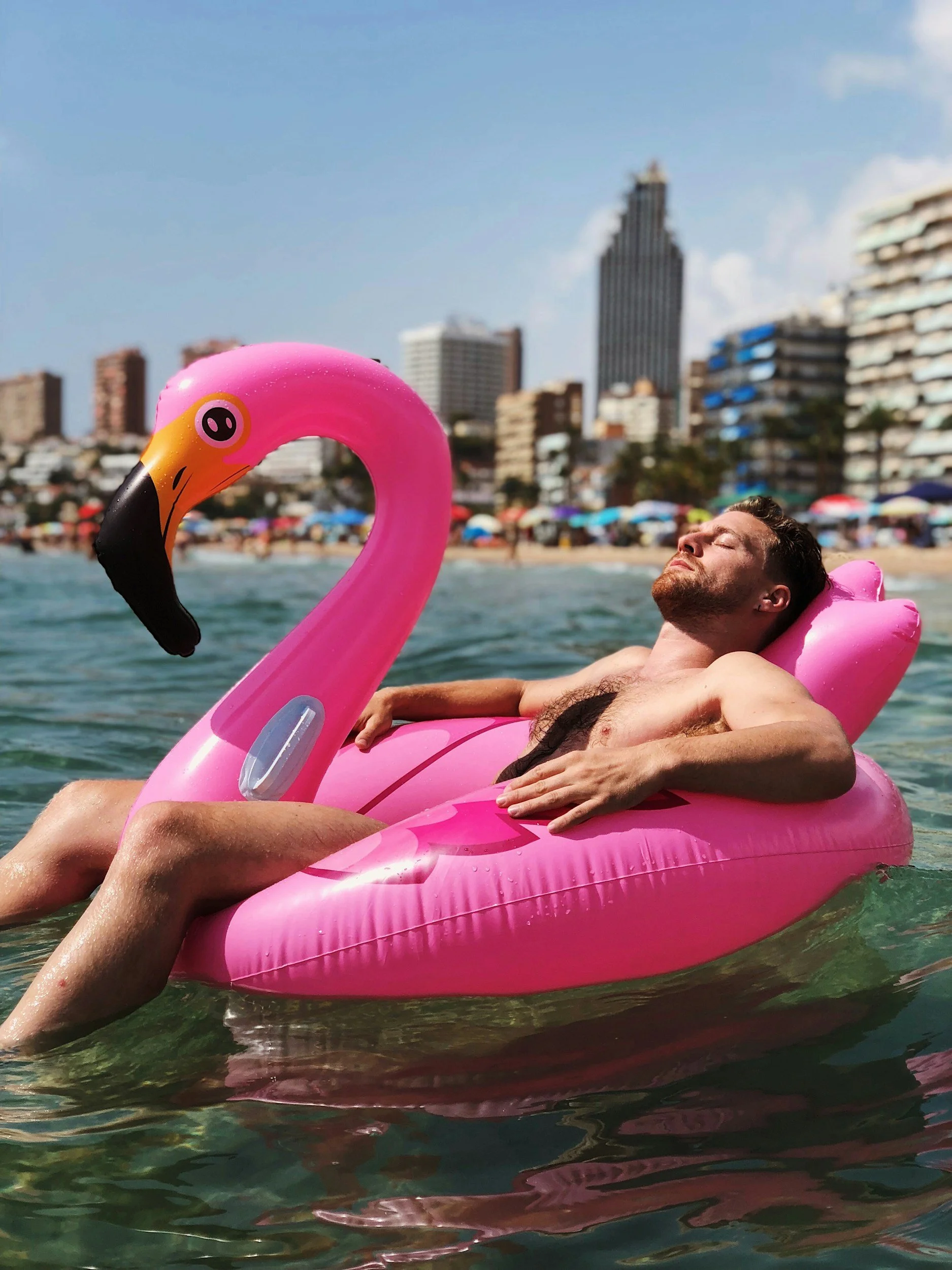 A man is relaxing on a pink flamingo-shaped inflatable float in the water at a beach, with city buildings and umbrellas in the background.