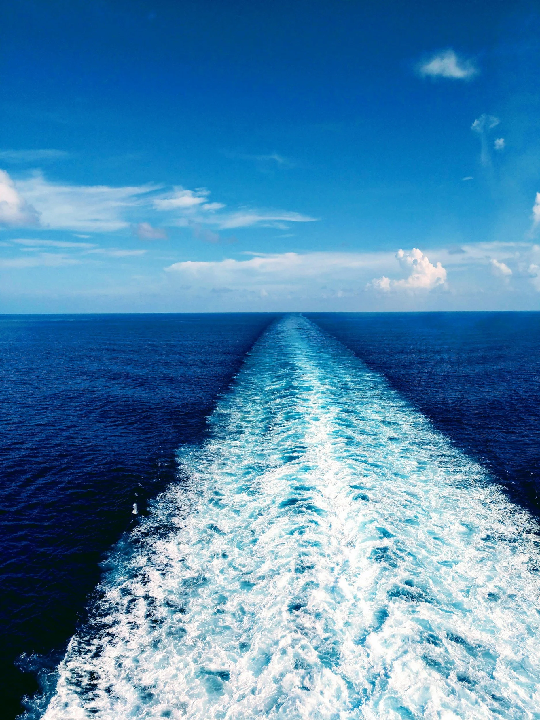View of the ocean with a boat trail in the water, under a blue sky with some clouds.