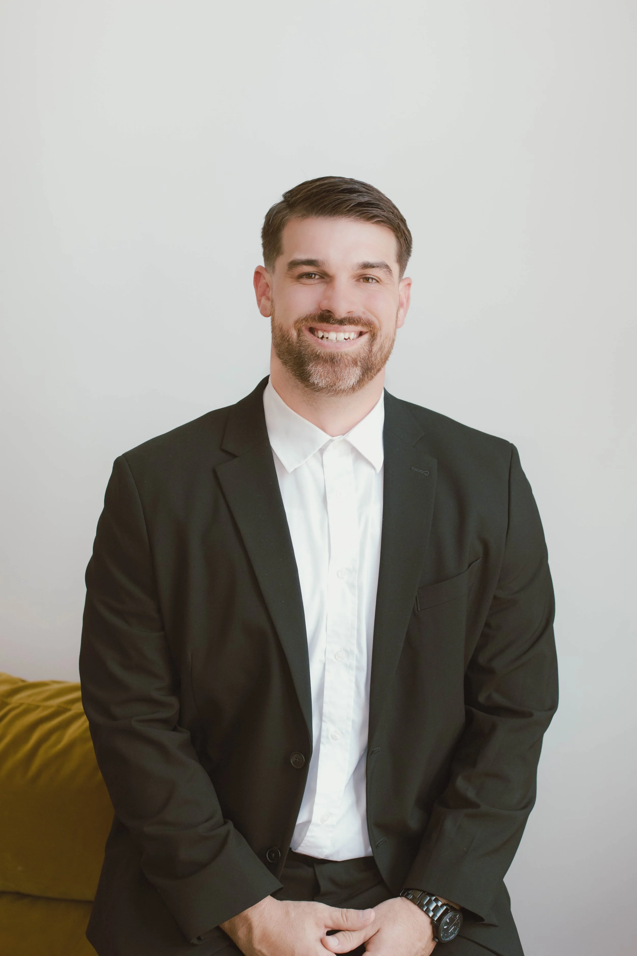 A man with brown hair and a beard wearing a black suit and white shirt, smiling and looking at the camera, standing in front of a plain white wall.