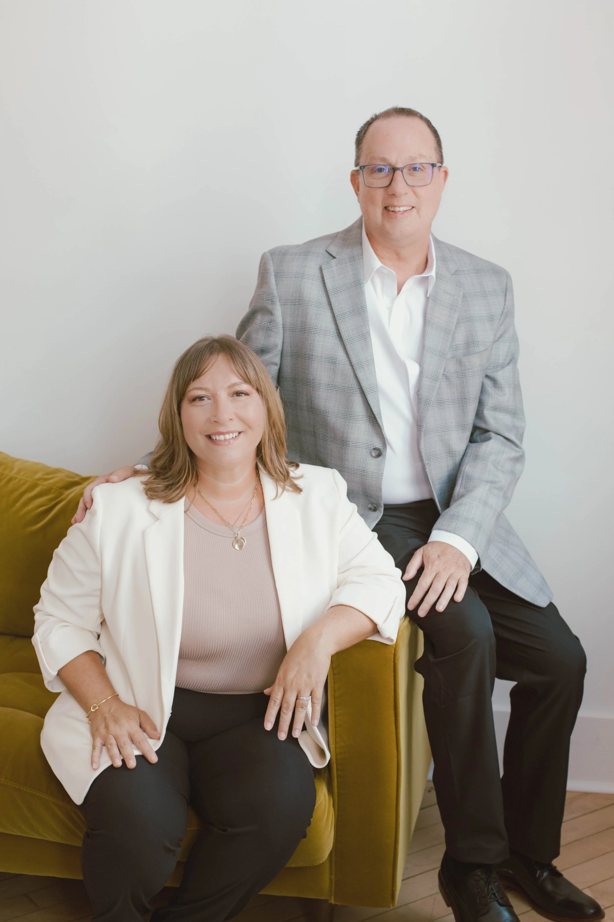 A man and a woman are posing for a photo in a room with white walls, with the man standing beside the woman who is seated on a mustard-colored sofa. The man is wearing glasses and a gray plaid blazer, while the woman is wearing a white blazer over a beige top and black pants.