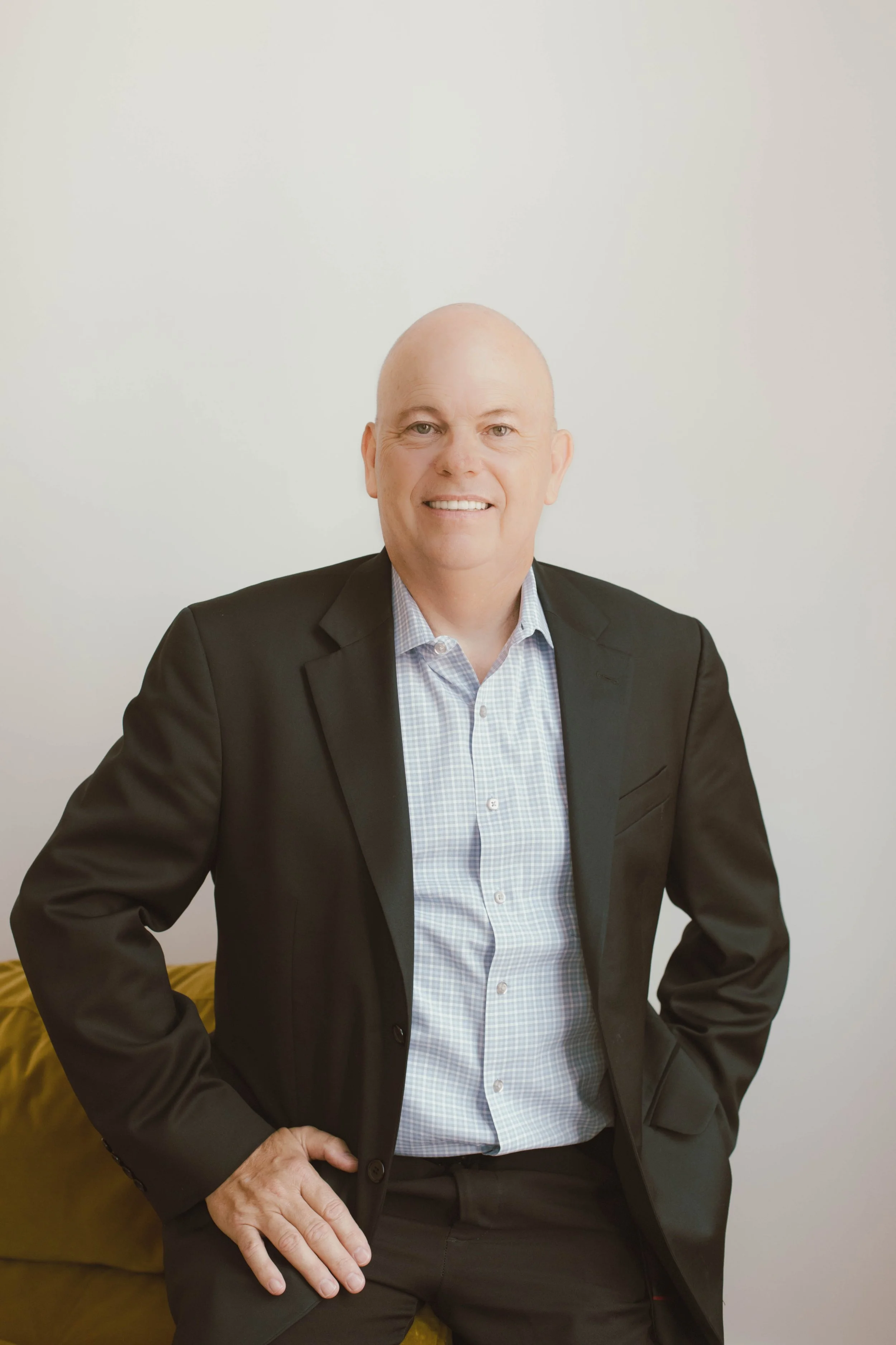 A bald man in a business suit smiling and standing with one hand on his hip in front of a blank, light-colored wall.