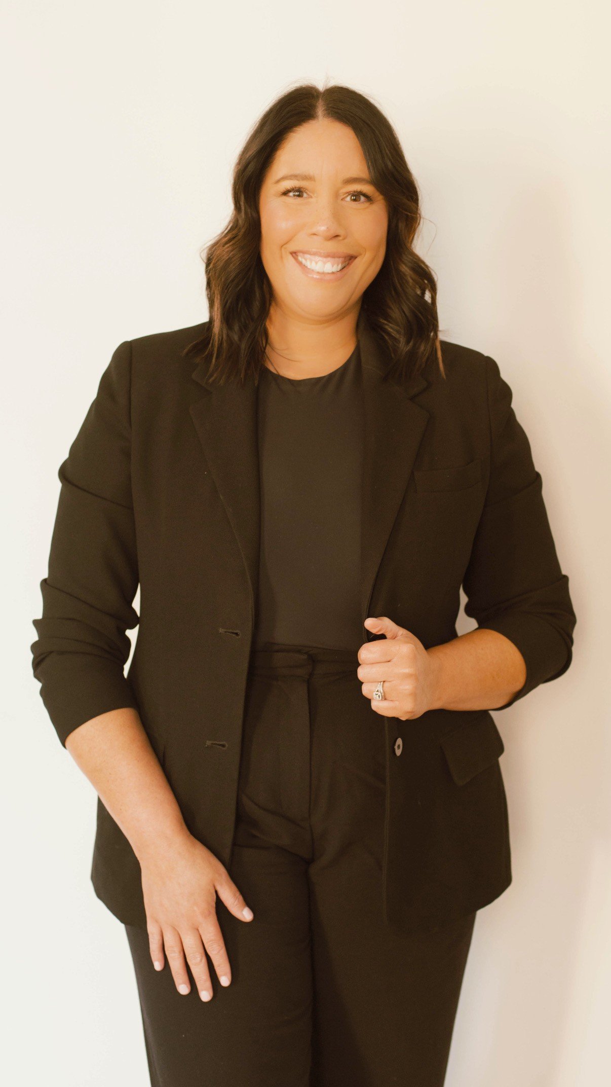 A woman with shoulder-length dark hair, smiling, wearing a black blazer and black top, sitting on a yellow couch with a plain light-colored wall in the background.