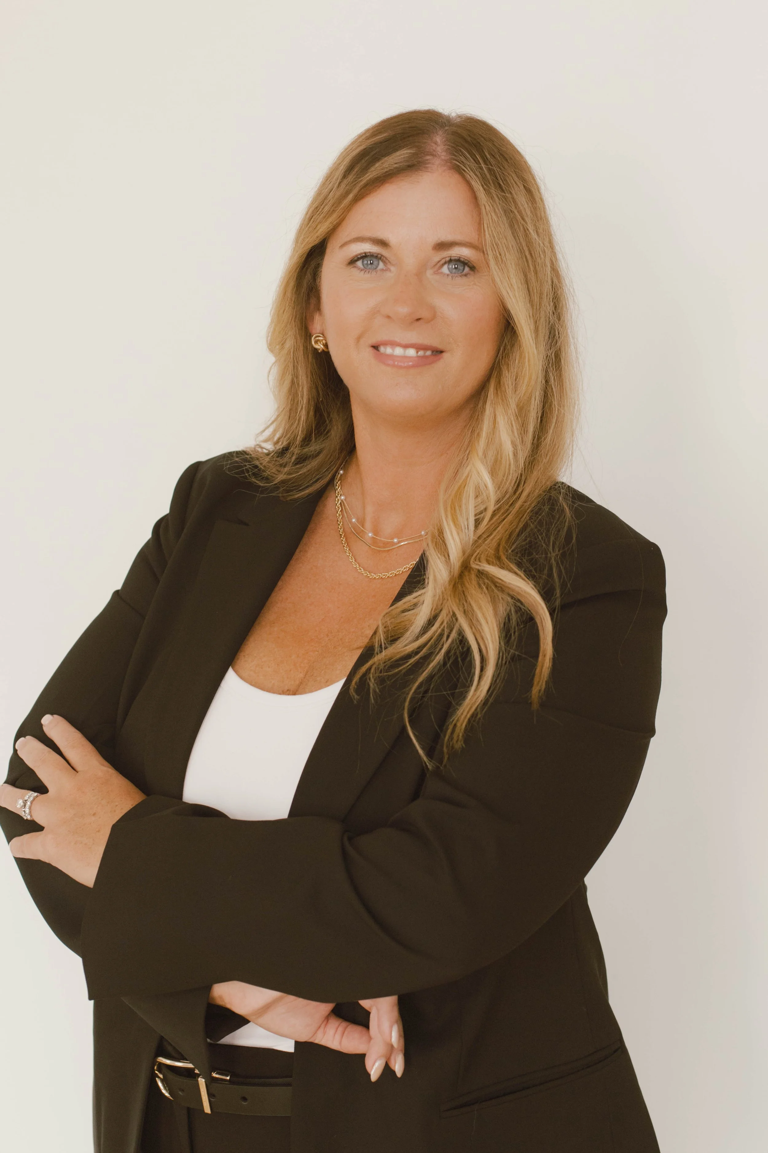 A woman with blonde hair styled in loose waves, wearing a black blazer over a white top, with gold jewelry, standing with arms crossed against a plain light background.