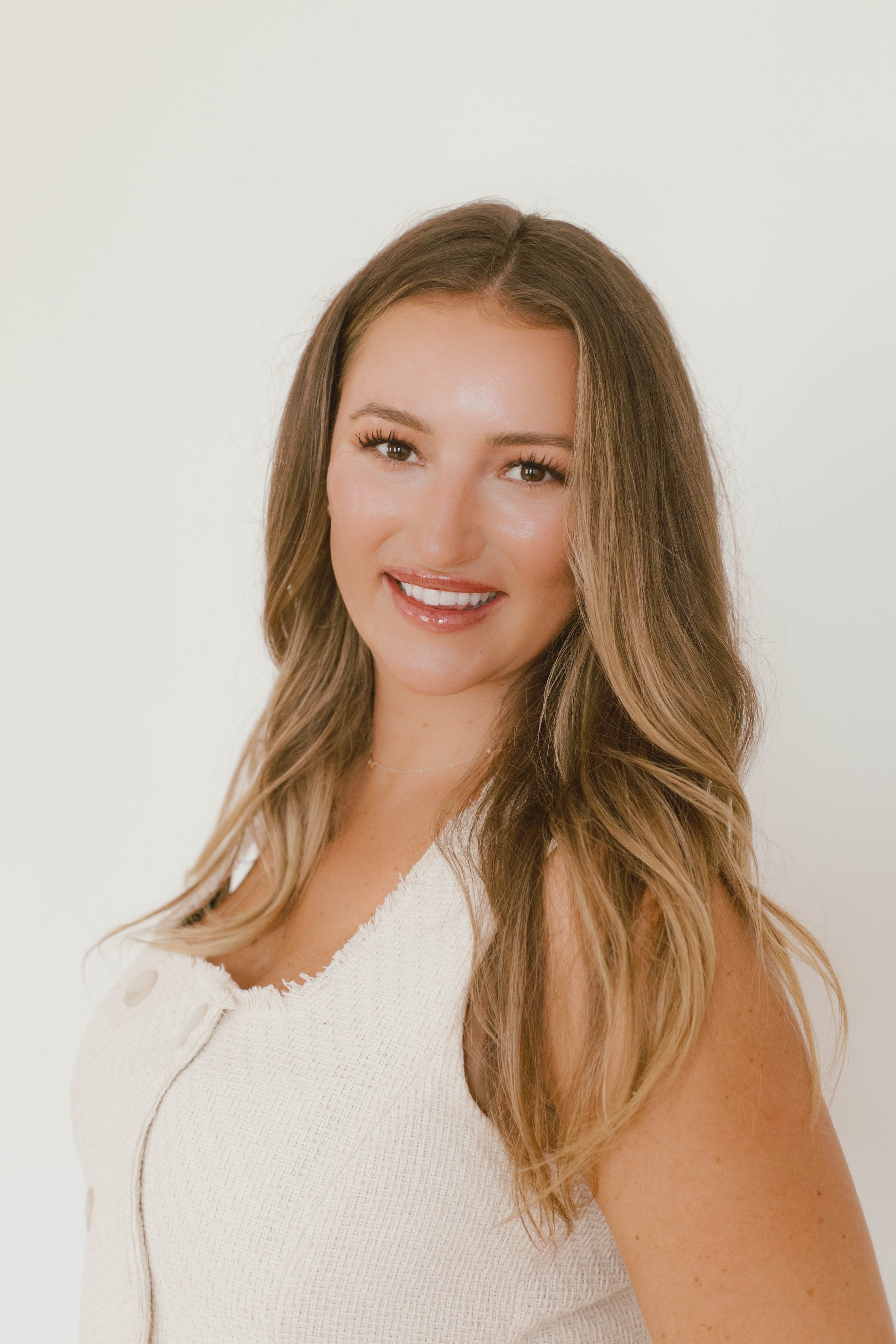 Portrait of a young woman with long, wavy blonde hair, smiling, wearing a sleeveless white top, against a plain white background.