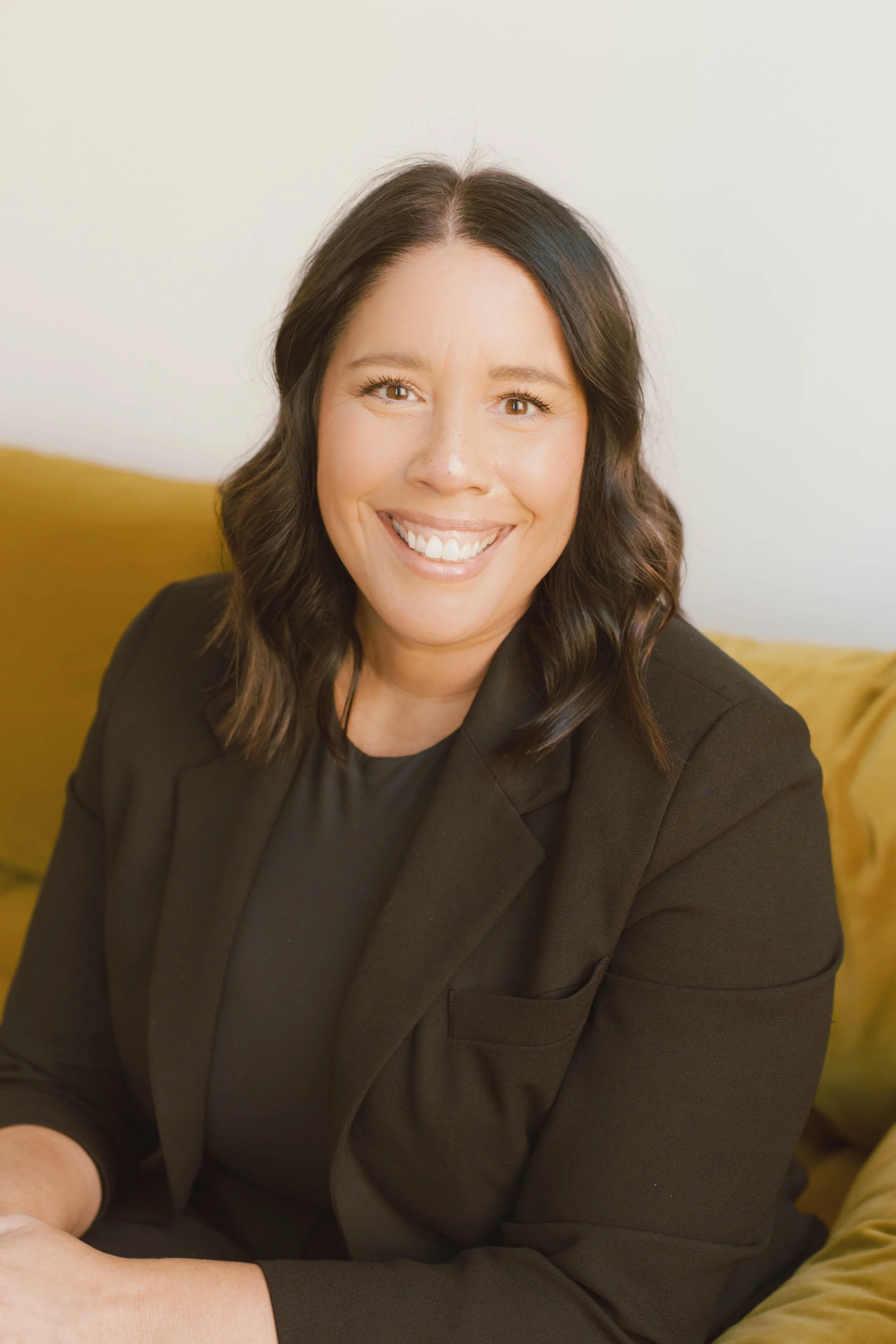 A woman with shoulder-length dark hair, smiling, wearing a black blazer and black top, sitting on a yellow couch with a plain light-colored wall in the background.