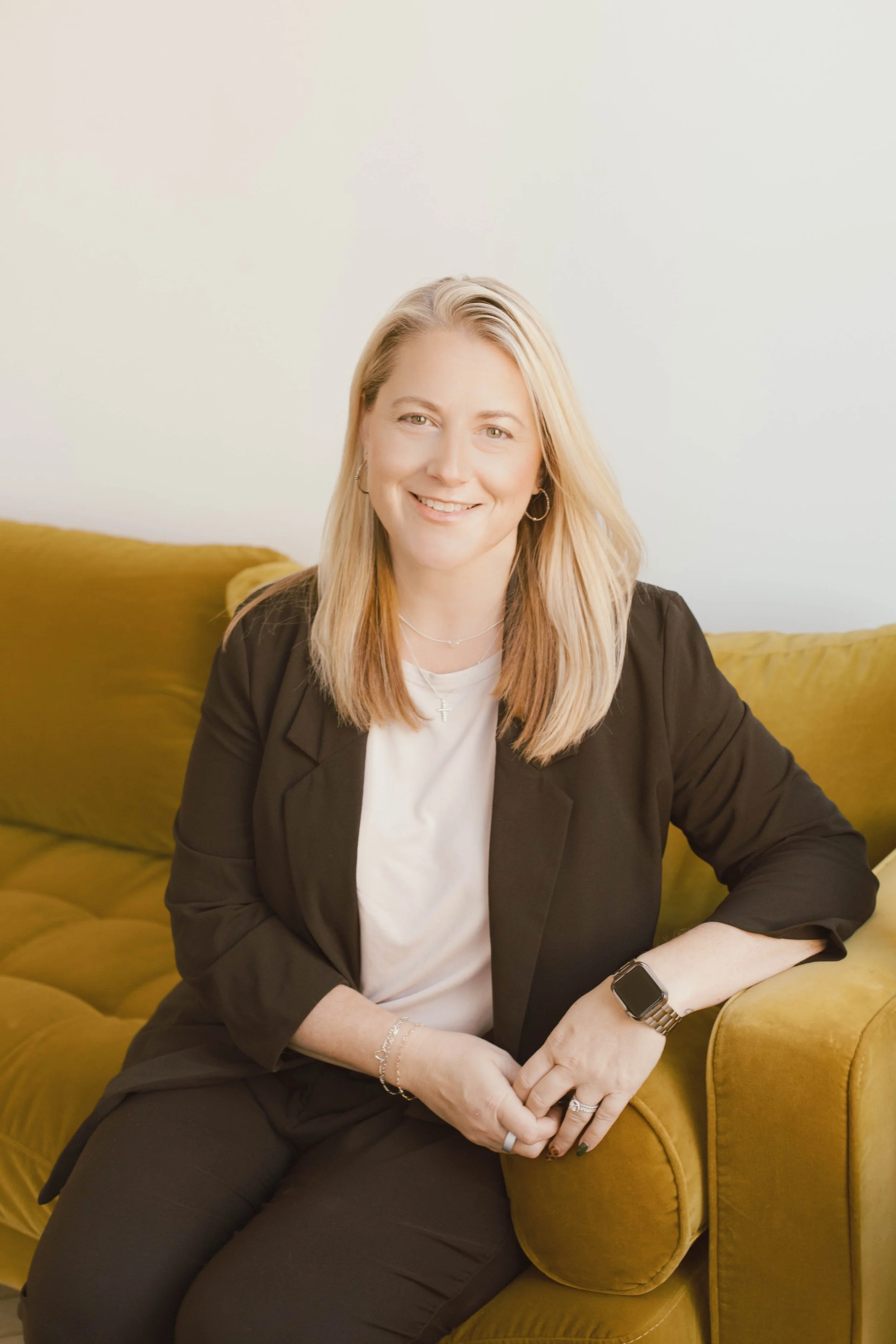 A woman with blonde hair smiling, sitting on a mustard yellow sofa, wearing a black blazer, white t-shirt, jewelry, and smartwatch.