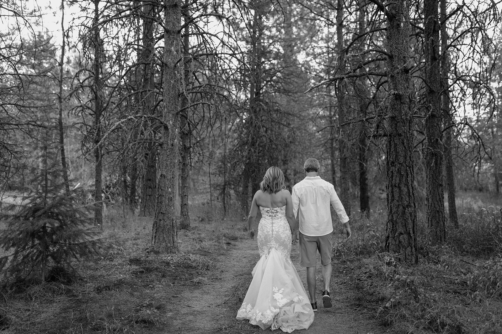 A bride and groom walking hand-in-hand along a trail in a forest, with tall trees surrounding them.