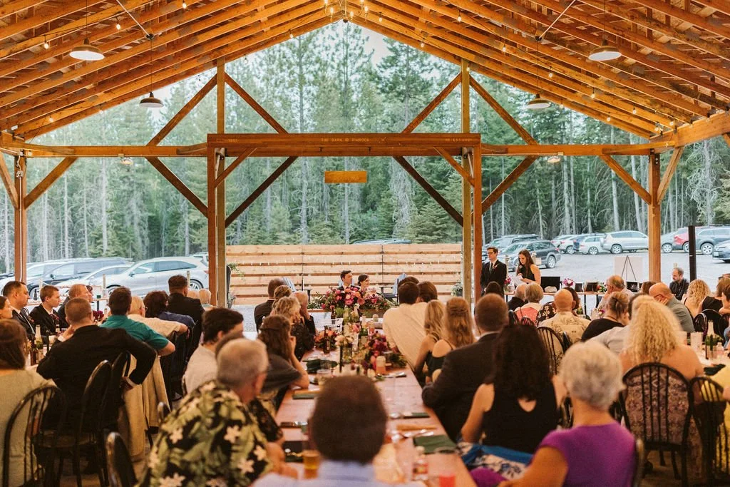 People seated at tables attending a wedding ceremony in a rustic wooden pavilion, with the bride and groom standing at the altar.