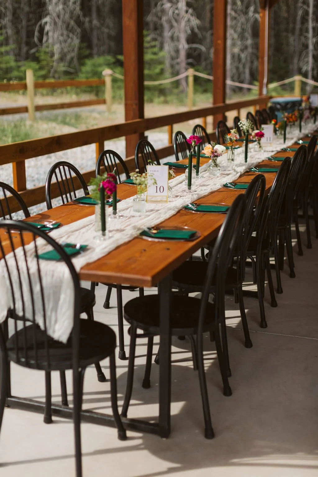 Long wooden table set with vases of pink and white flowers, green napkins, and a white table runner, surrounded by black chairs, outdoors with a forest background.