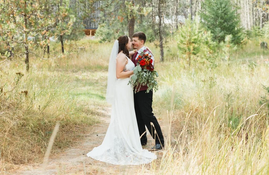 A bride and groom stand close together on a dirt path in a wooded area, smiling at each other; the bride wears a white wedding dress with a veil, and the groom holds a large bouquet of red and orange flowers with greenery.
