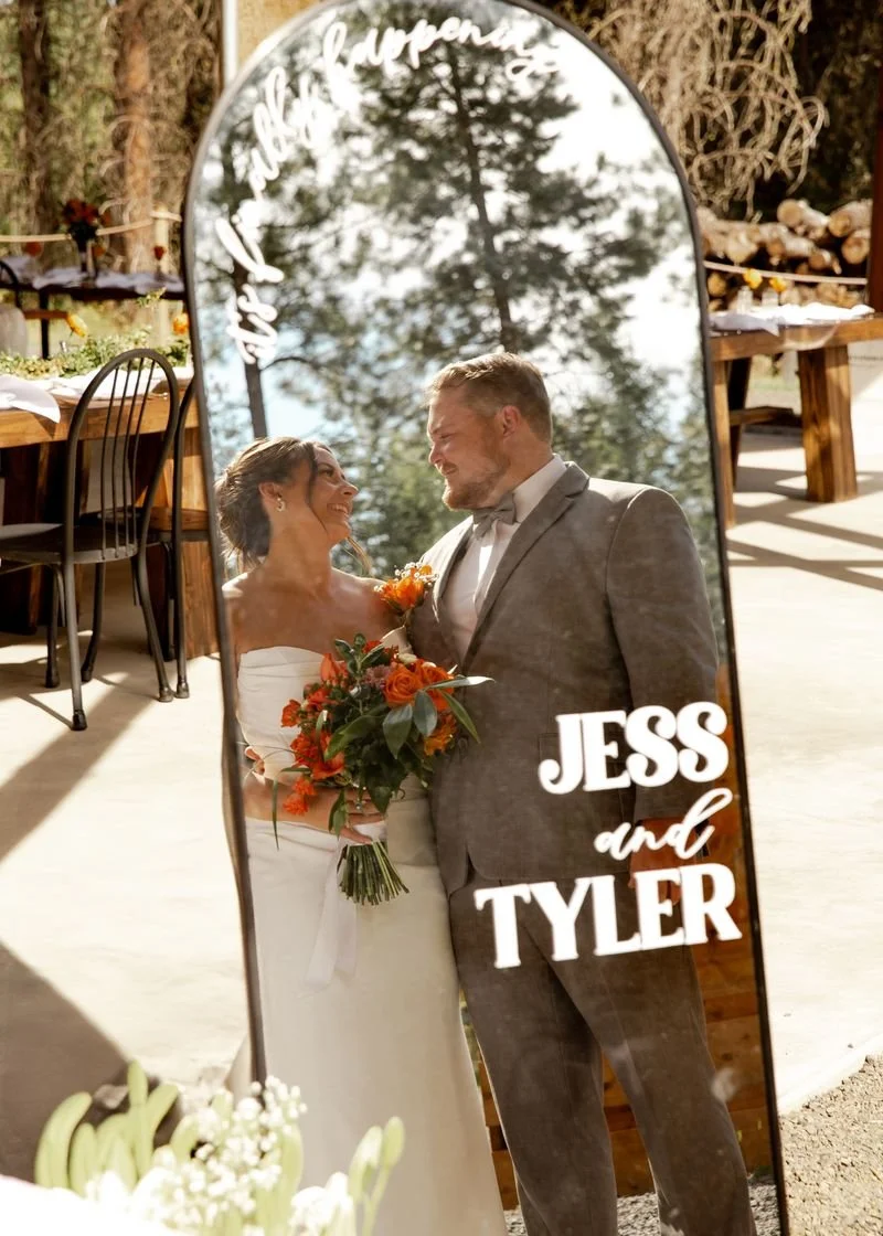 A bride and groom look at each other through a mirror with the words "JESS and TYLER" and a decorative phrase on it, at an outdoor wedding reception with trees and decorated tables in the background.