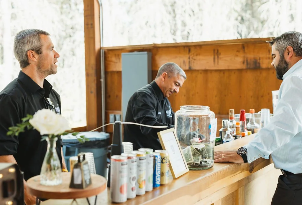 Two men are at a bar counter; one man is smiling and the bartender is handling cash and drinks, with a jar filled with money on the counter.