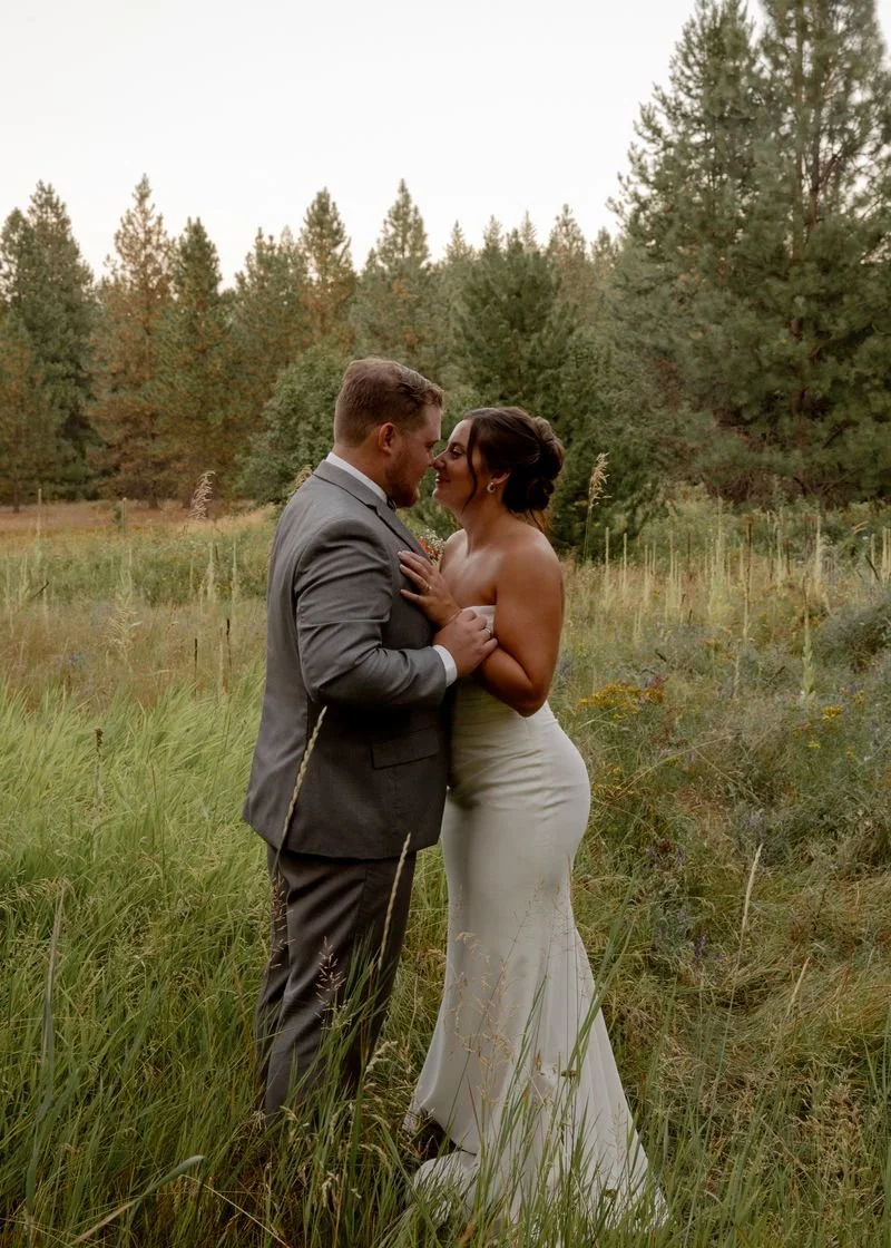 A couple dressed in wedding attire standing in a grassy field with trees in the background, leaning close with noses touching and hands on each other's chest.