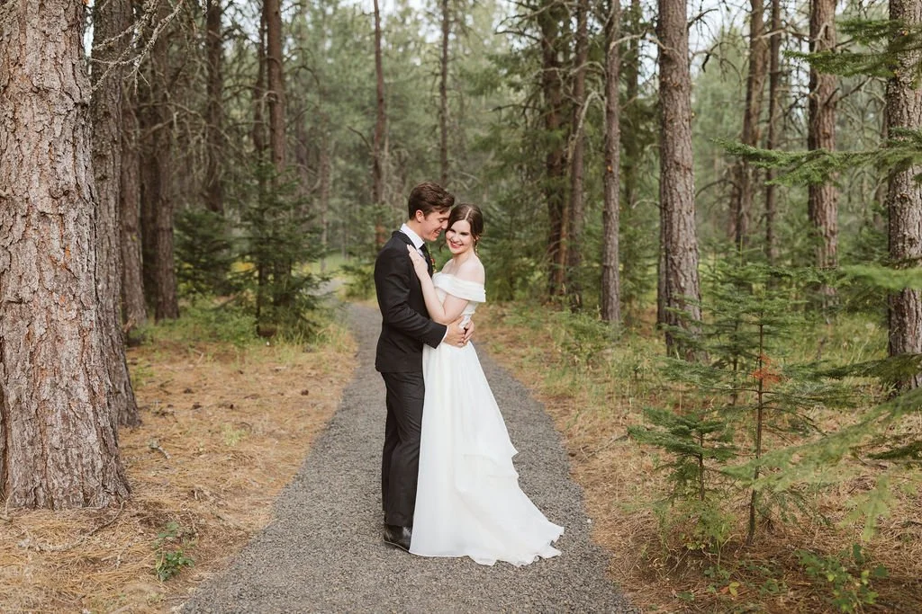 A newlywed couple in wedding attire embracing and smiling in a forested area.