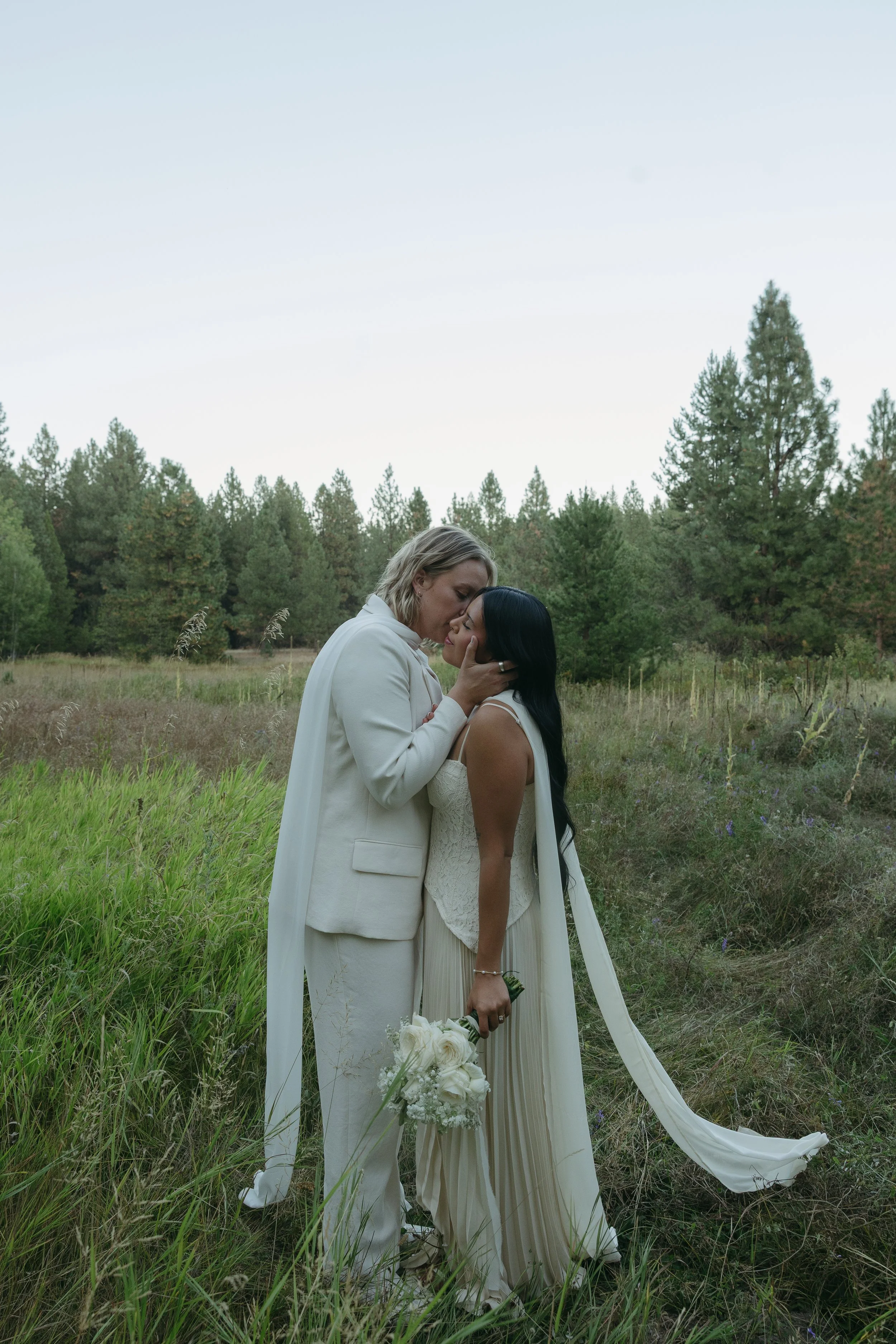 Two women in white wedding attire sharing a kiss in a grassy outdoor setting with trees in the background.