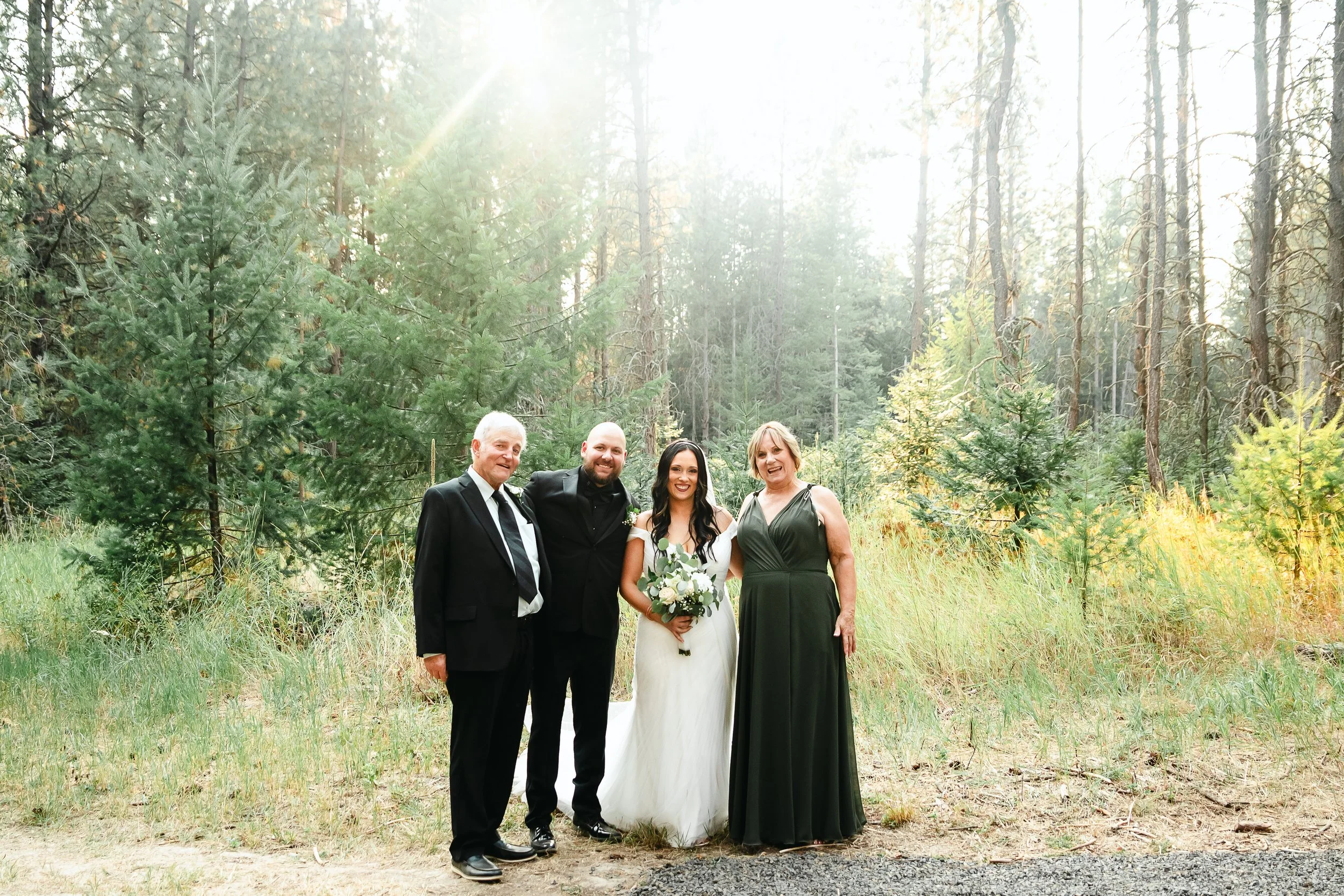 A group of four people at a wedding outdoors in a forest with tall trees and sunlight shining through.