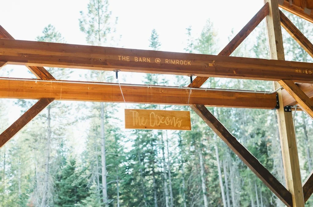 A partially built wooden outdoor structure with a sign hanging from it, surrounded by tall trees in a forest setting.