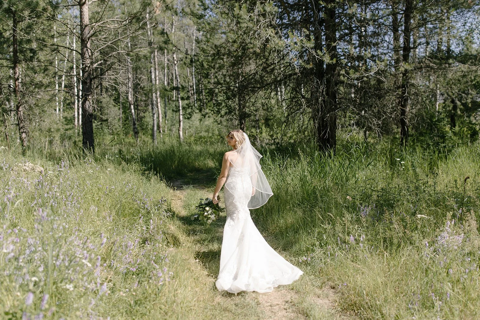 A bride in a white wedding dress and veil walking on a narrow dirt path through a green forest, holding a bouquet of flowers.