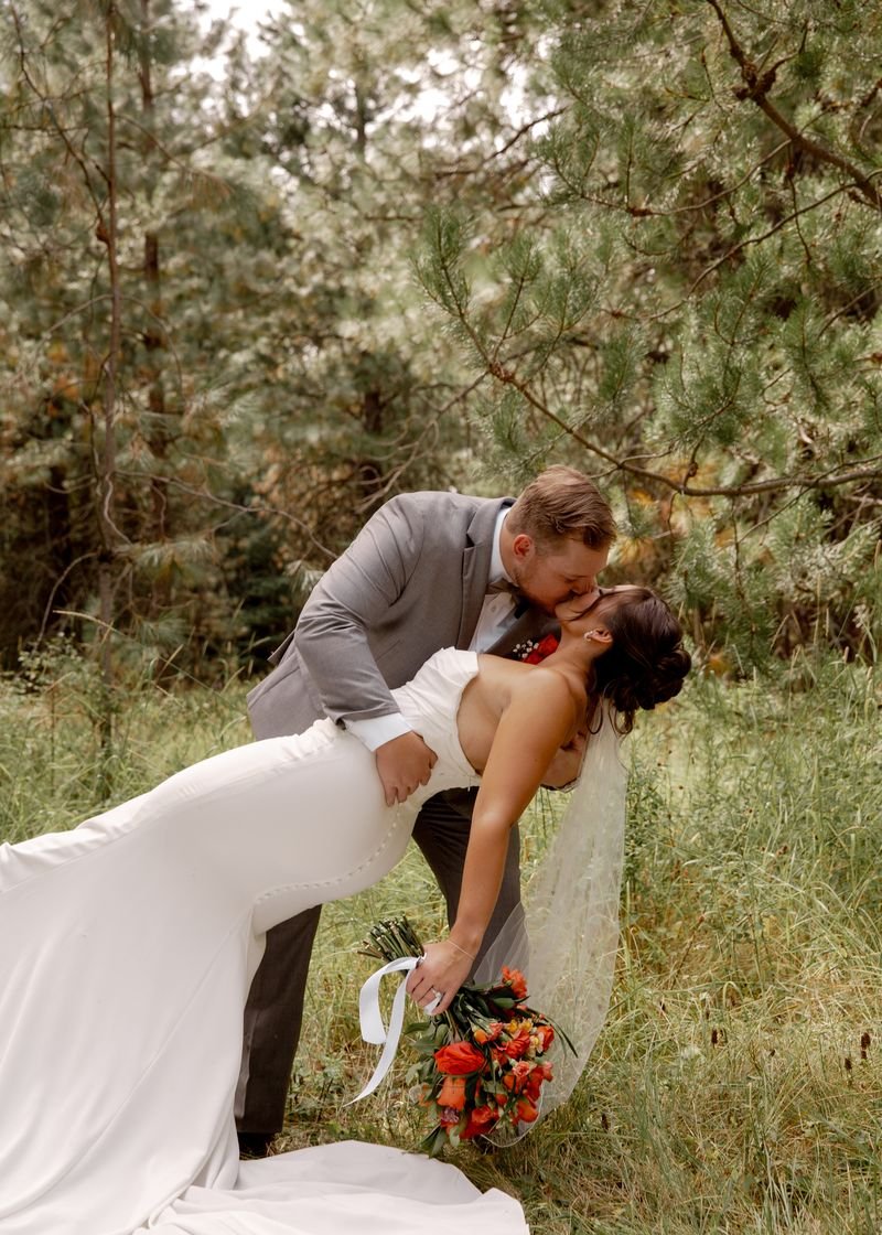 A newlywed couple shares a kiss in a forest, with the groom dipping the bride. The bride is holding a bouquet of orange flowers and wearing a strapless white wedding dress with a veil. The groom is dressed in a light gray suit.