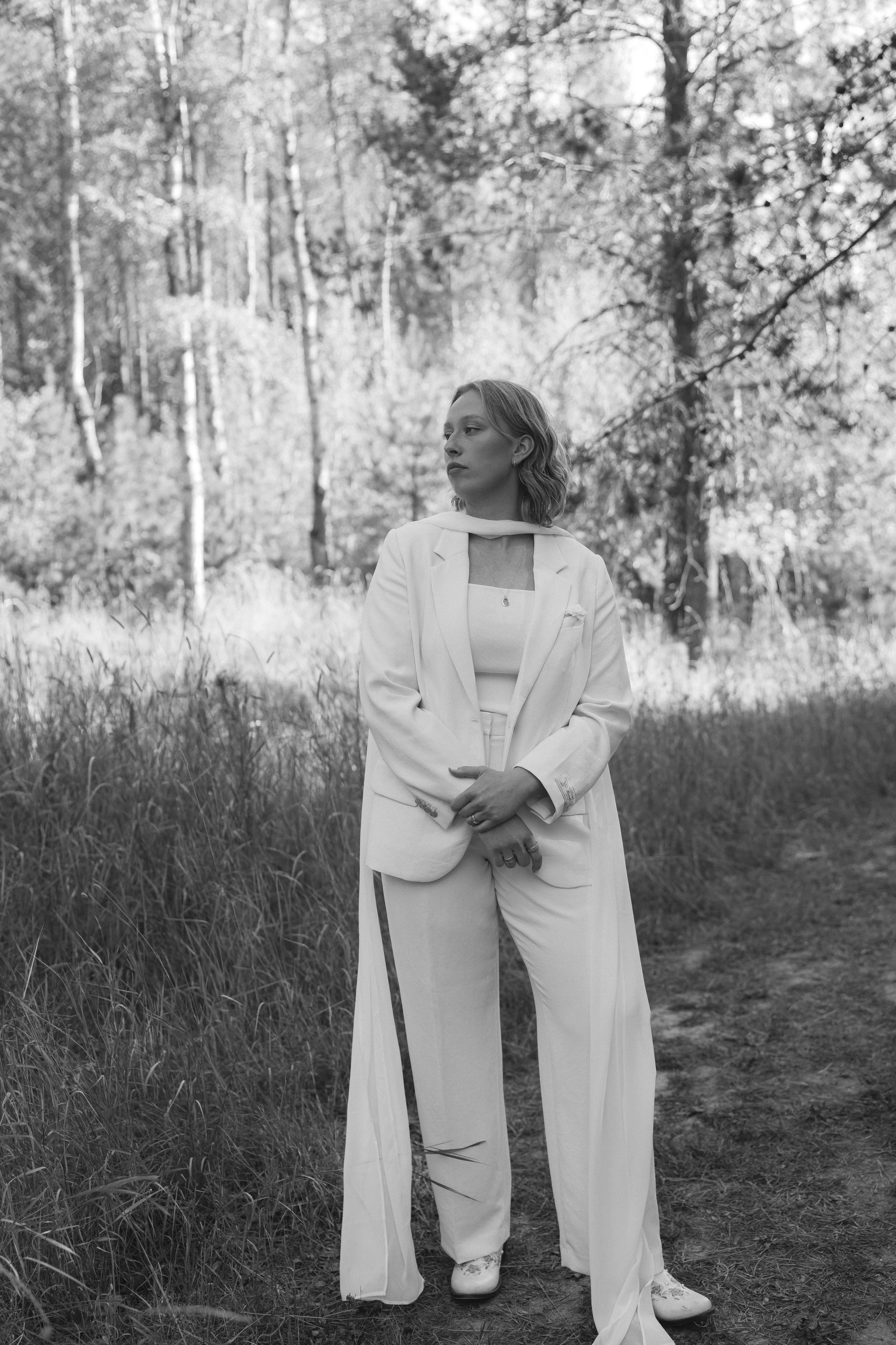 A woman dressed in a white suit and shoes standing outdoors in a wooded area with tall grass, looking to the side.