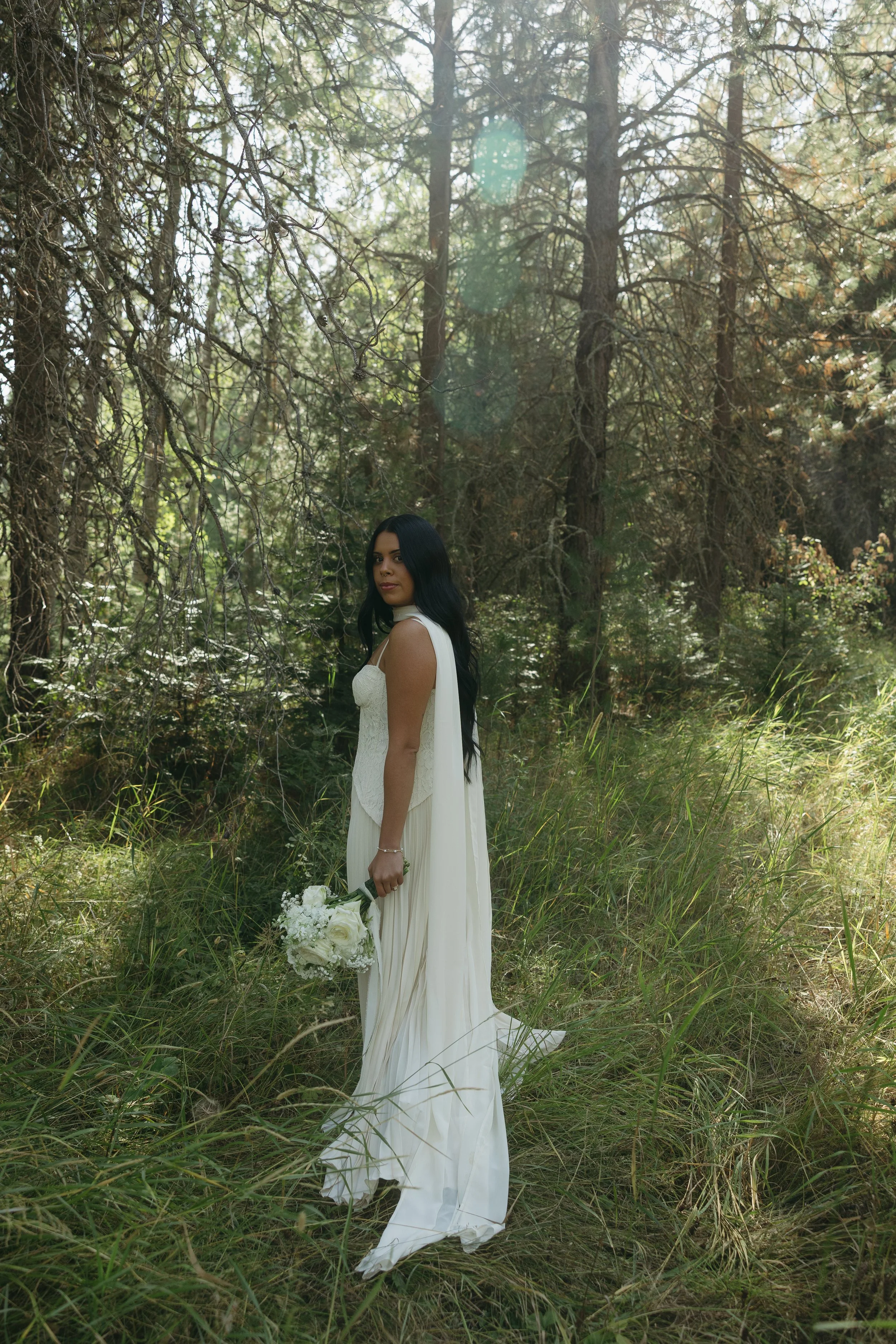 A woman in a white dress holding a bouquet of white flowers standing in a forest with tall grass and trees.