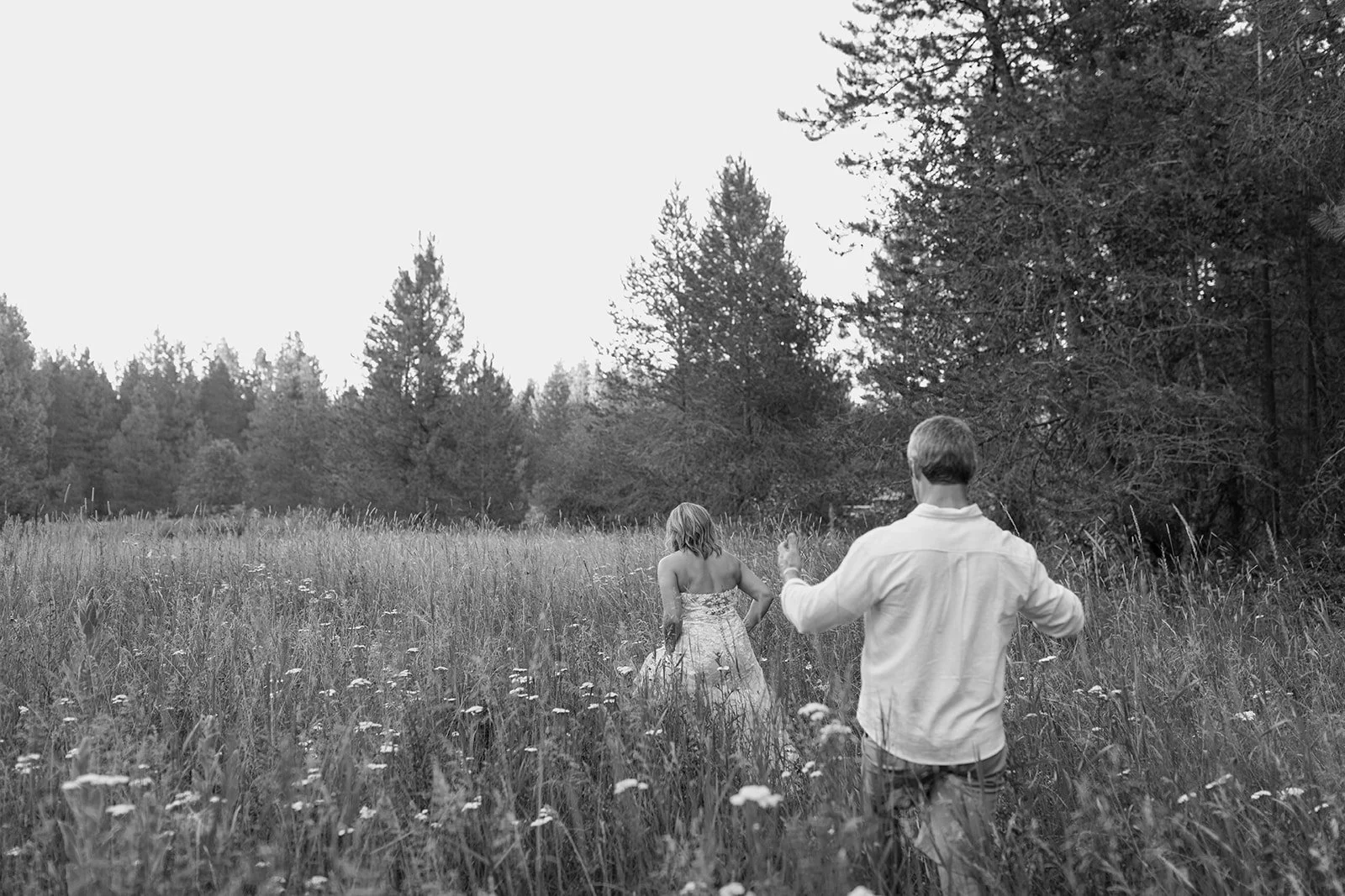 A boy and girl walking through a grassy field with tall wildflowers, with trees in the background.