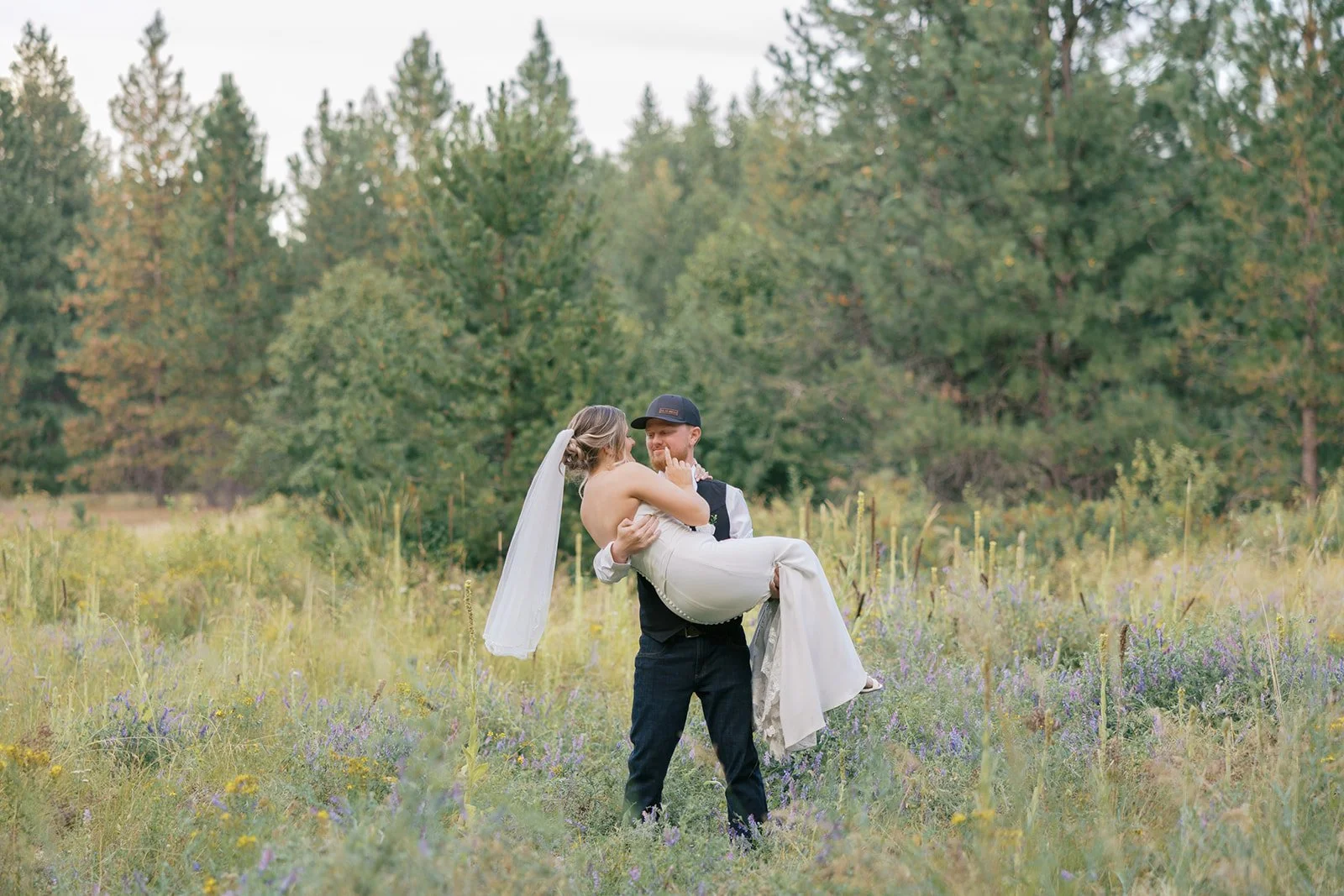 A groom in a black vest and baseball cap holding a bride in a white wedding dress and veil in a field with green trees in the background.