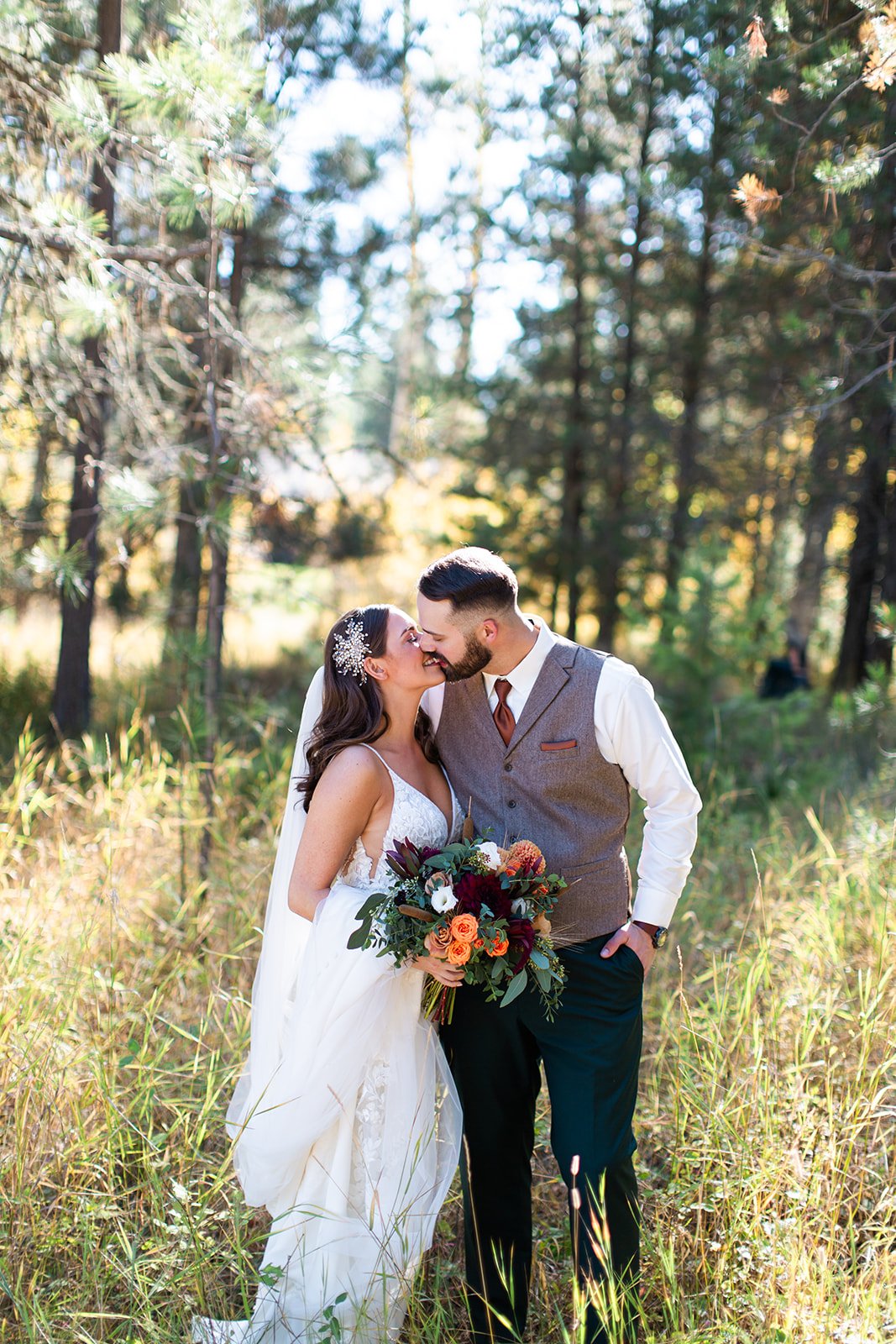 A bride and groom sharing a kiss in a forest setting during their wedding. The bride is wearing a white wedding dress with lace details and a floral hairpiece, holding a bouquet of flowers. The groom is dressed in a gray vest and white shirt, with hi