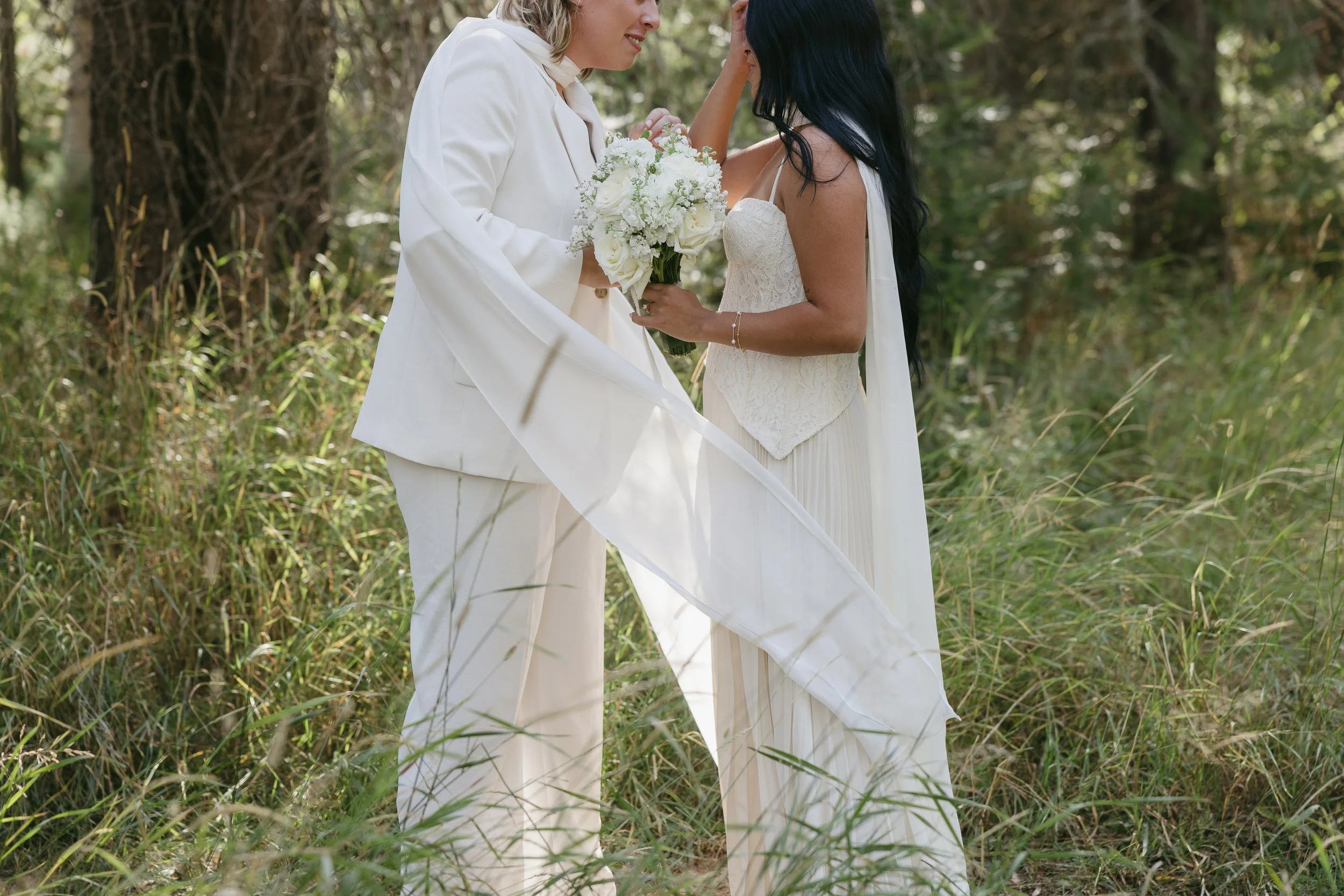 Two women in white attire standing closely in a natural outdoor setting, sharing a tender moment. One woman holds a bouquet of white flowers.
