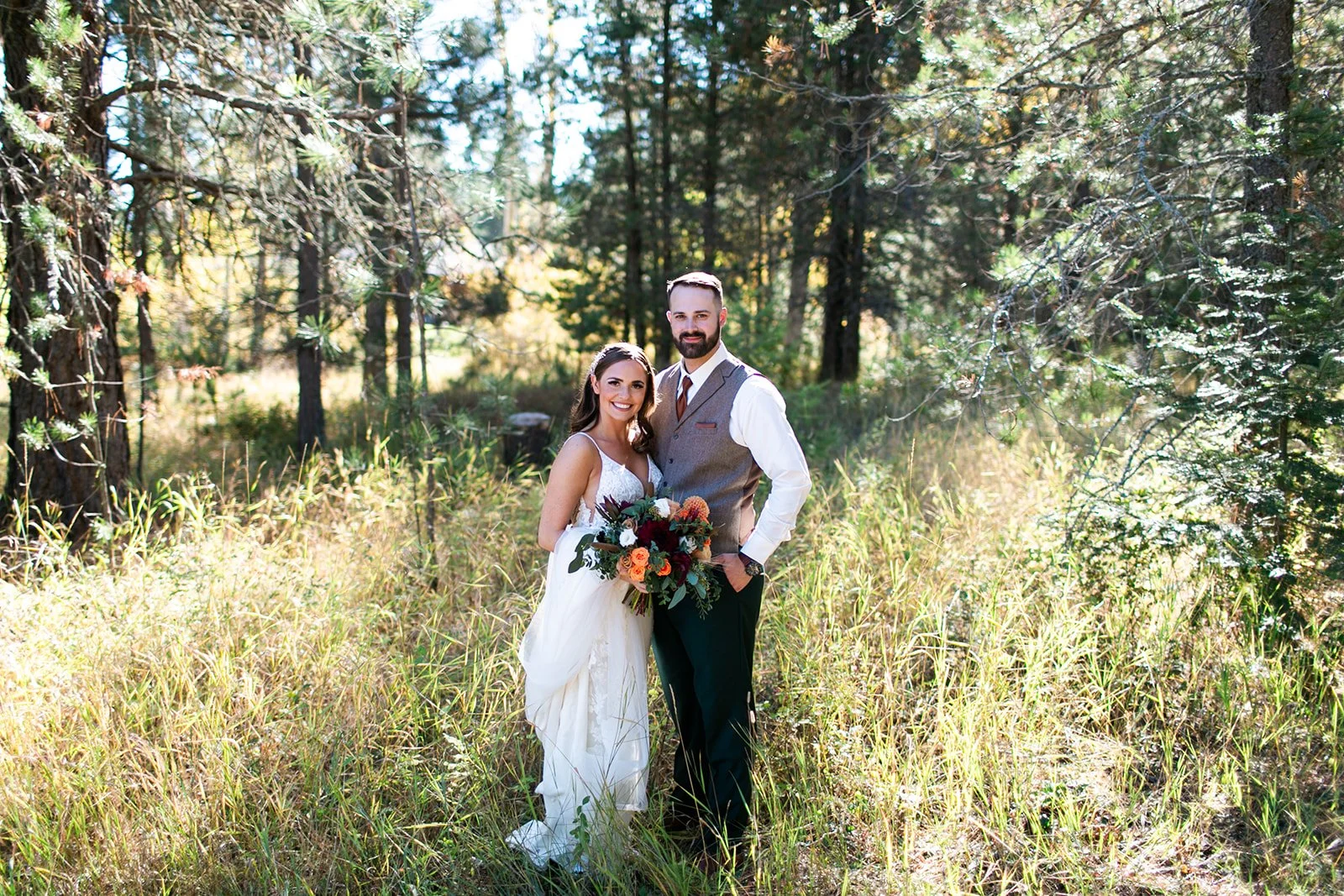A couple in wedding attire standing in a wooded outdoor setting with sunlight filtering through trees, holding a bouquet of flowers.