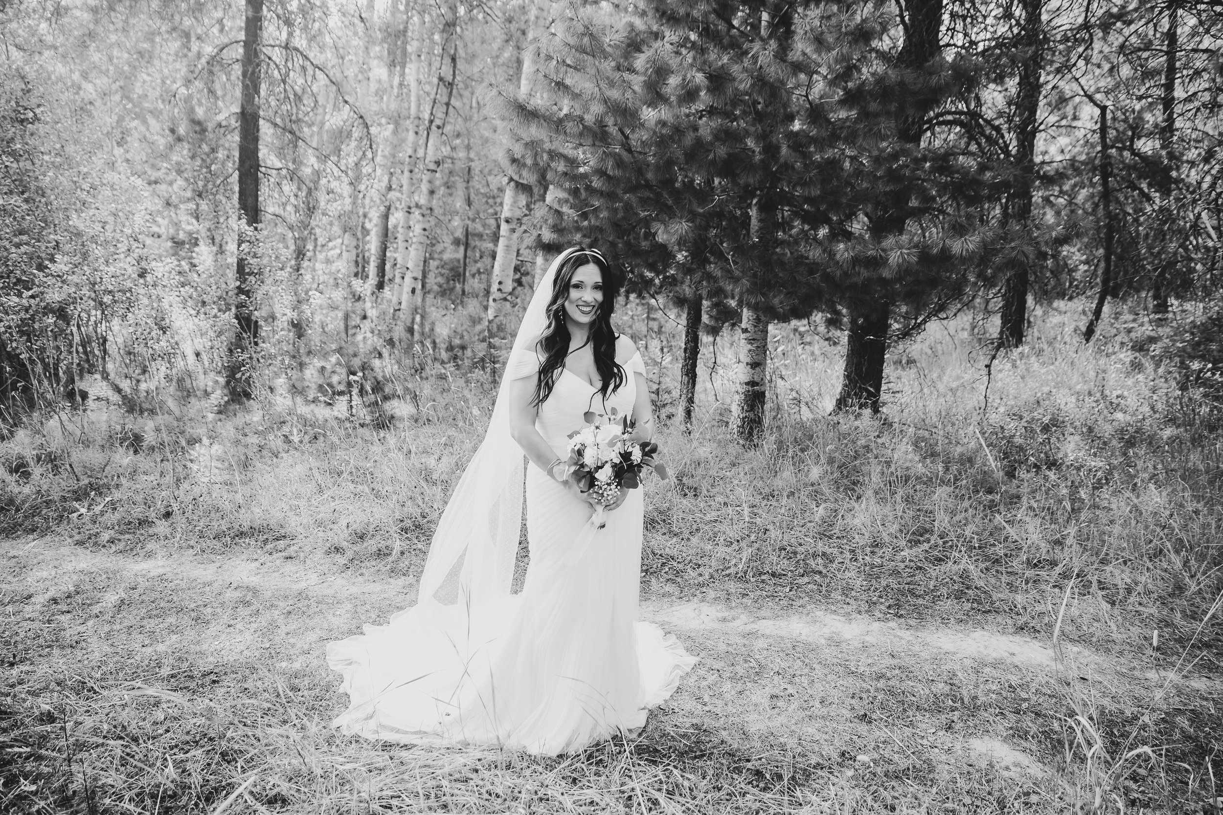 Black and white photo of a smiling bride in a wedding dress holding a bouquet, standing on a forest trail with trees and grass in the background.