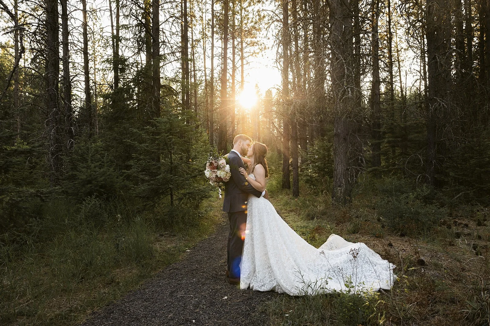 A bride and groom embrace in a forest trail at sunset, with the sun shining behind trees, the bride holding a bouquet, and the bride wearing a white wedding gown.