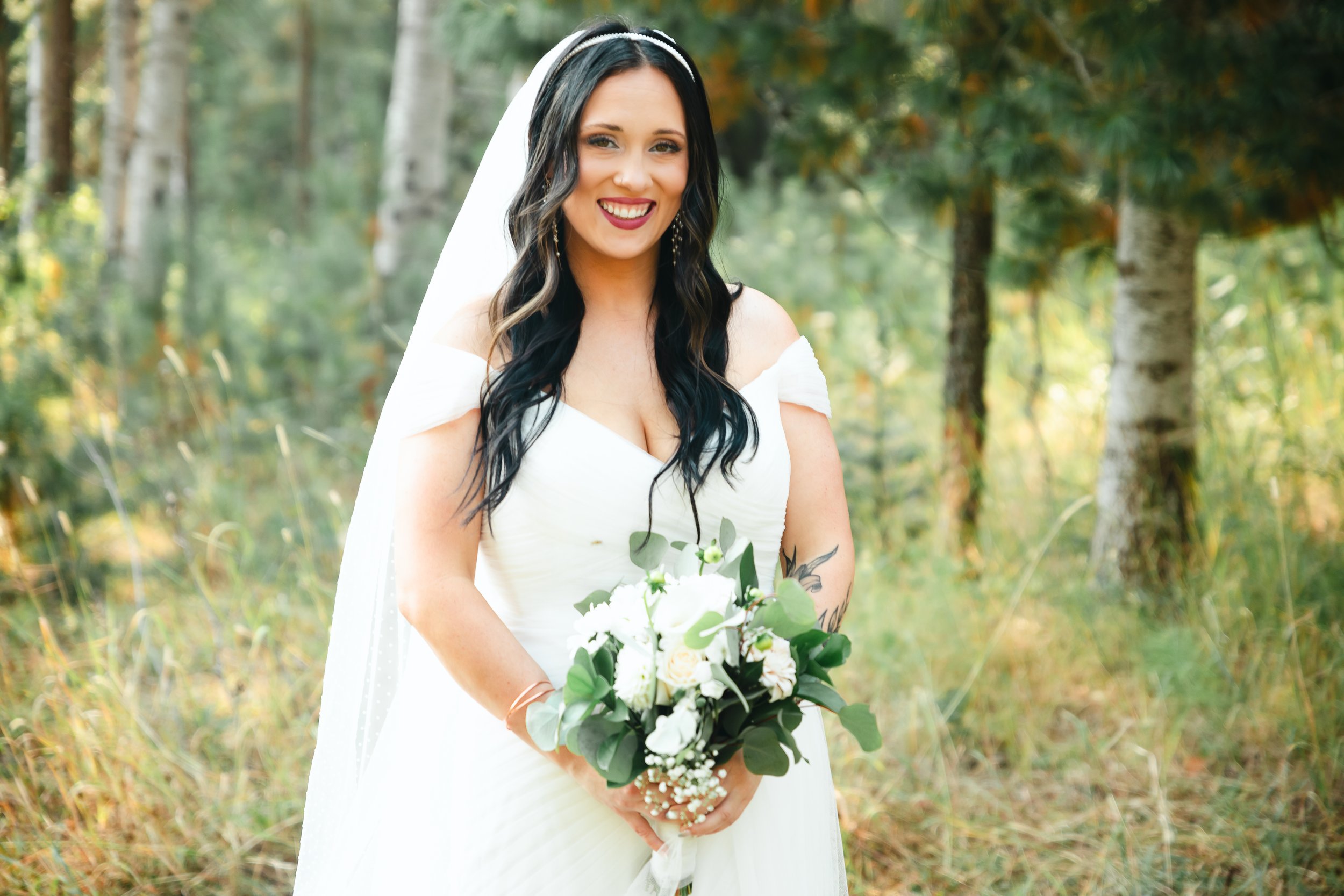 A woman in a white wedding dress holding a bouquet of white and green flowers, standing outdoors in a wooded area.