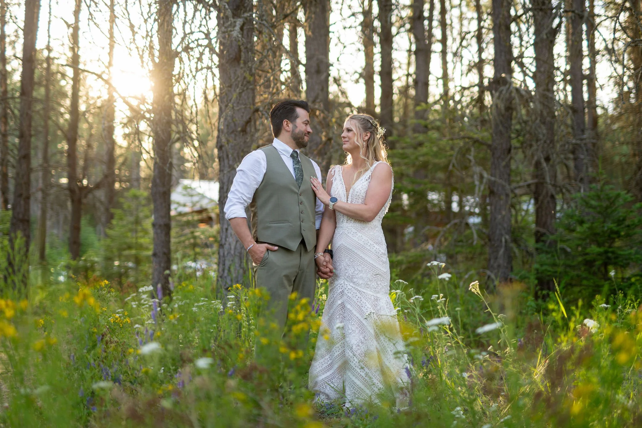 A couple in wedding attire stands holding hands in a forest with tall trees, wildflowers, and sunlight filtering through the branches.