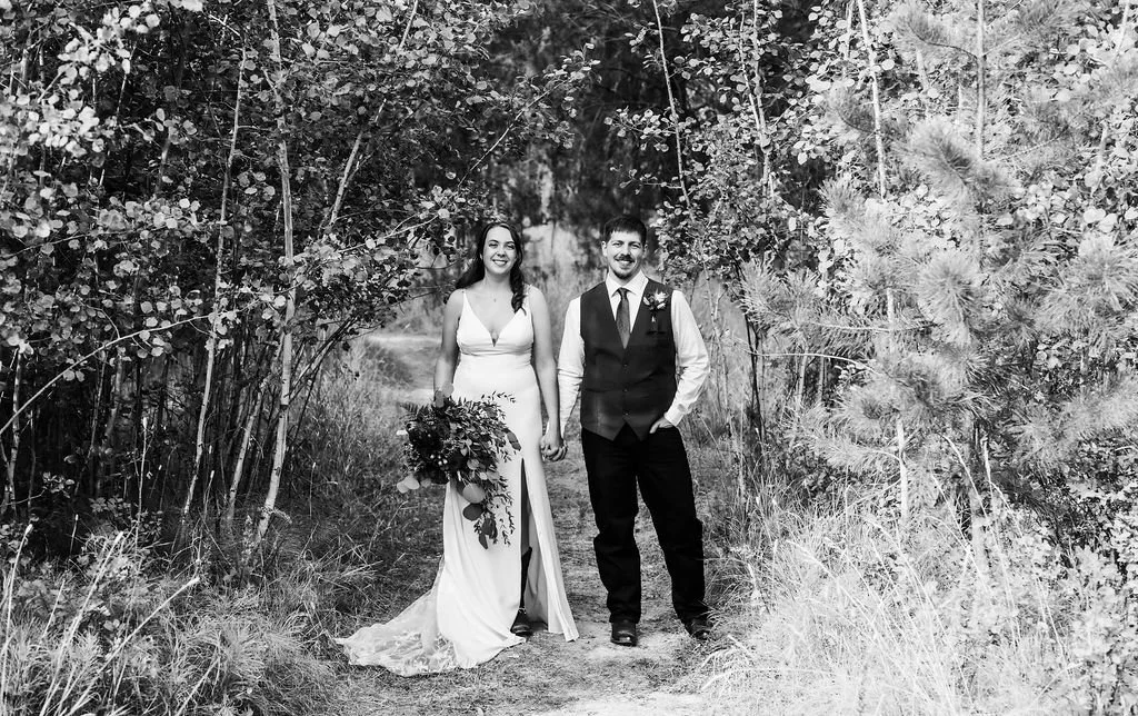 A bride and groom holding hands on a trail surrounded by trees, smiling. The bride wears a white wedding dress and holds a bouquet, while the groom wears a vest, tie, and dress shirt.