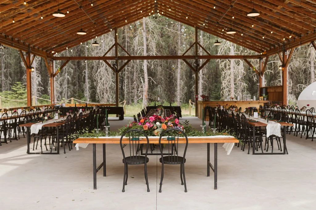 Indoor event space with long tables, black chairs, floral centerpiece, string lights, and a wooden ceiling with open sides revealing trees outside.