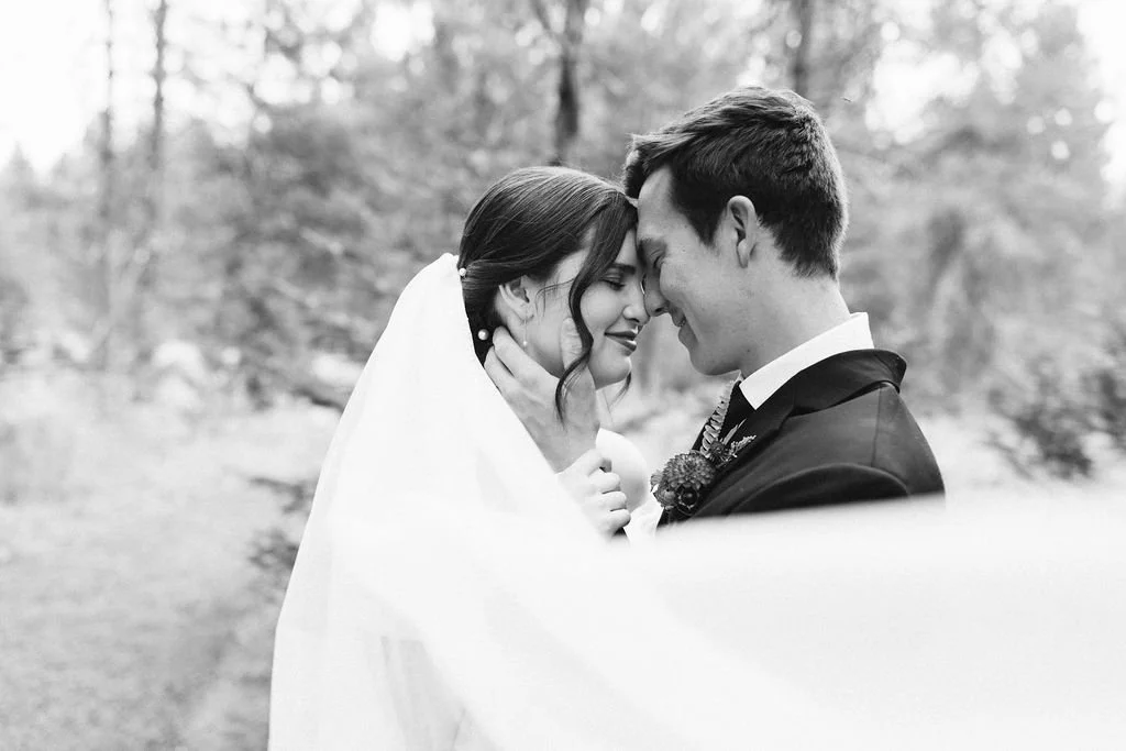 A black and white photo of a newlywed couple with foreheads touching, smiling, outdoors in a wooded area.