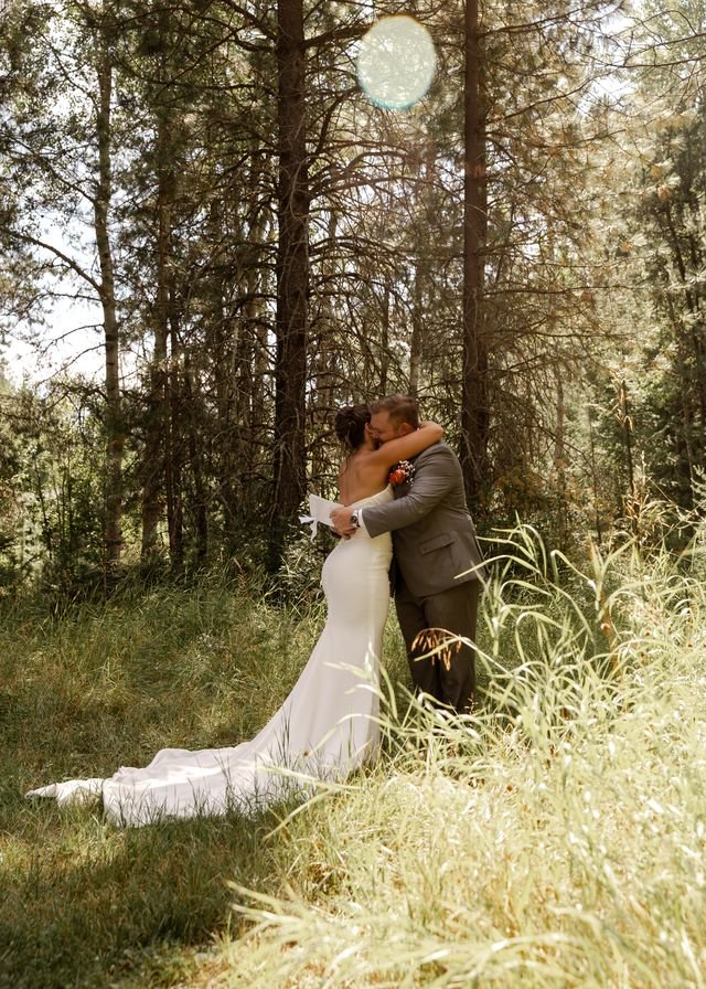 A bride and groom embracing outside in a wooded area with sunlight filtering through the trees.