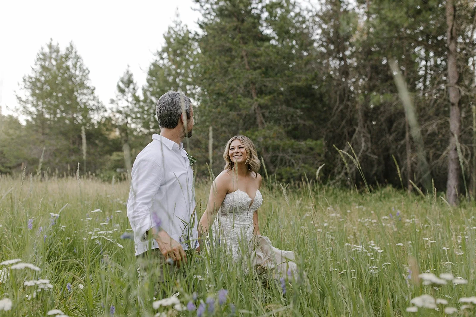 A man and woman holding hands and walking through a grassy field with wildflowers and trees in the background.