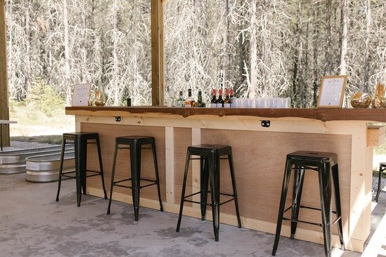 A wooden outdoor bar with four black stools, bottles and glasses on the counter, and framed signs in a rustic setting with forest in the background.