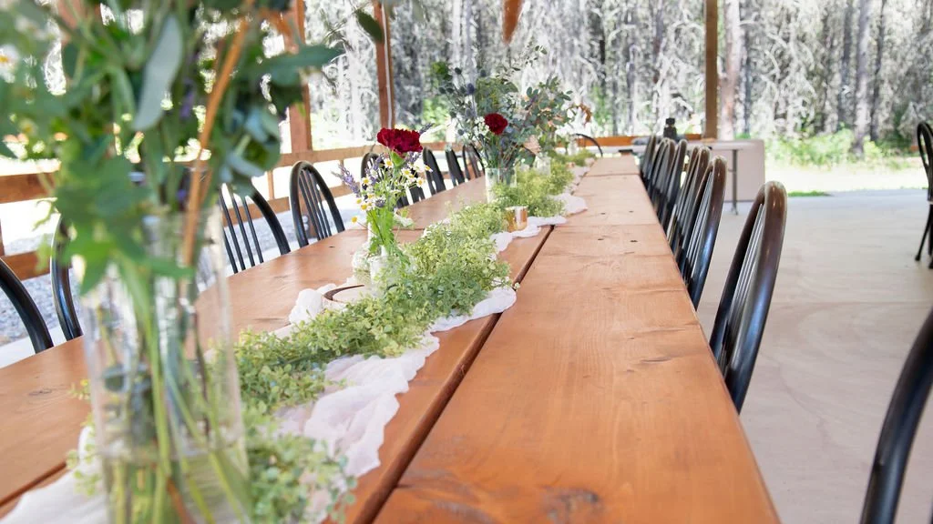 Long wooden table set with vases of red and purple flowers and greenery in a rustic outdoor setting with trees in the background.