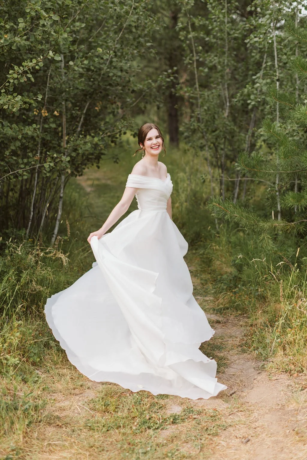 A woman in a white wedding dress standing on a dirt path in a green forest, smiling and holding the side of her dress.
