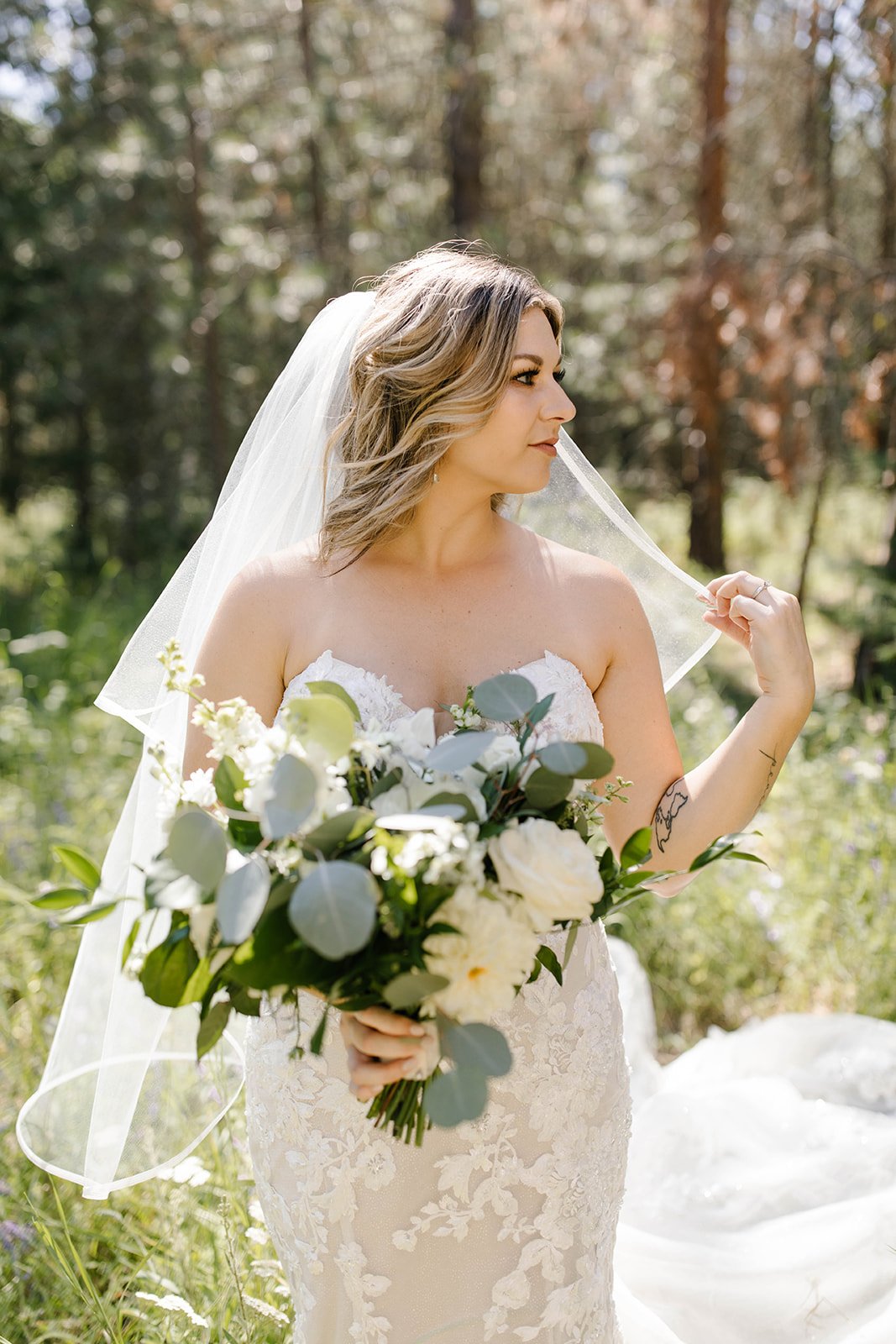 A bride in a white lace wedding dress holding a bouquet of white roses, eucalyptus, and greenery, standing outdoors in a sunlit forest with trees in the background.