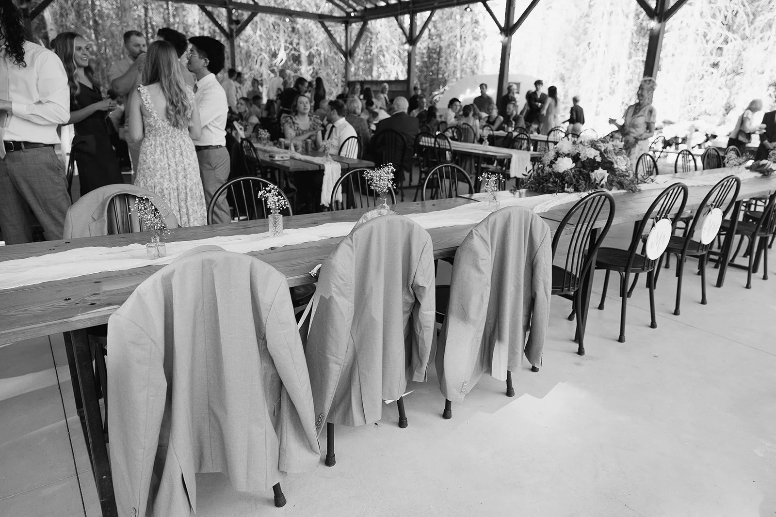 A wedding reception with a long decorated table in the foreground, featuring flower arrangements and coat jackets hanging on chairs. In the background, guests are mingling and sitting at tables, with a rustic, open-sided venue and trees visible outsi