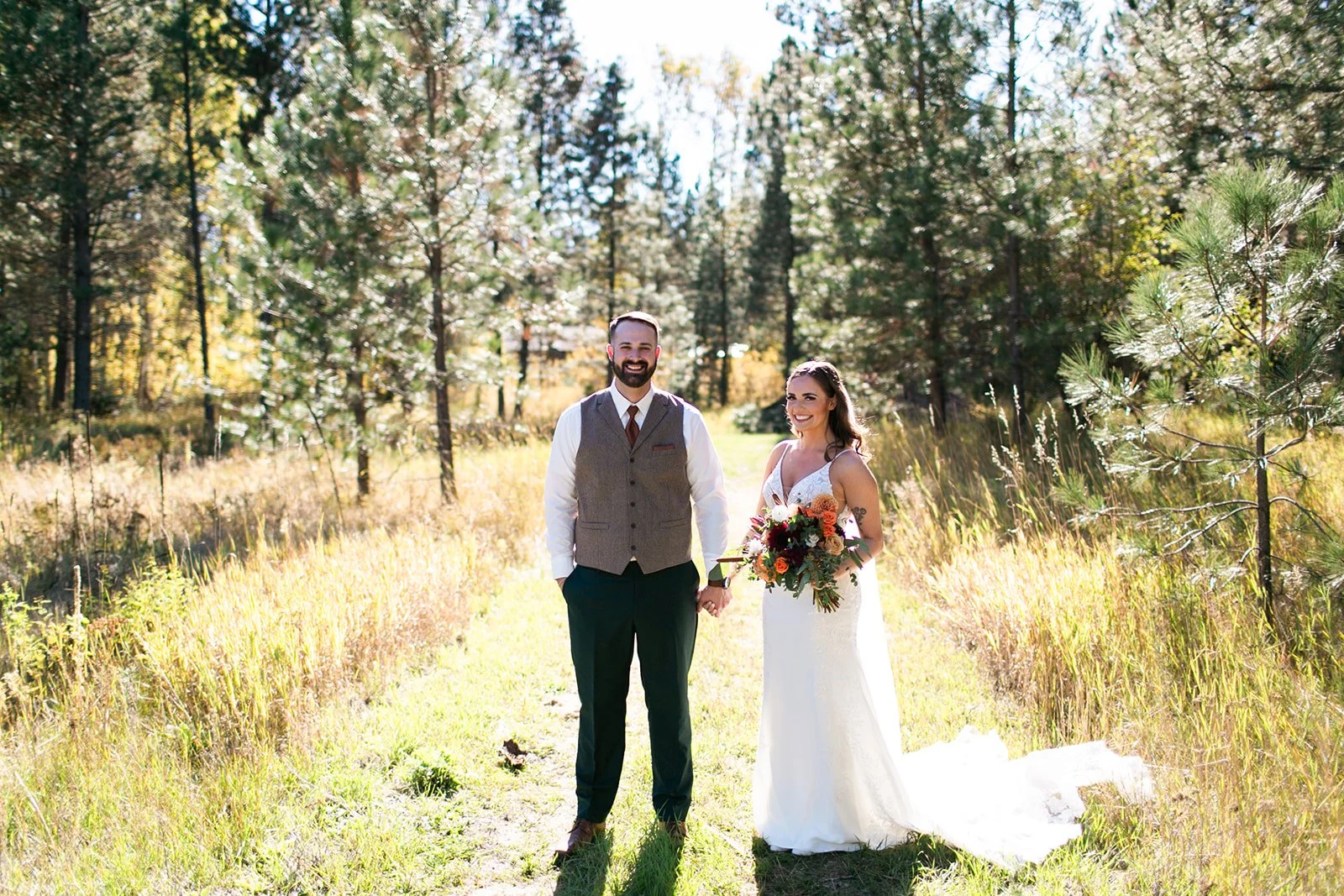 A newlywed couple holding hands and smiling in a forested outdoor setting during daytime, with trees and sunlight in the background.