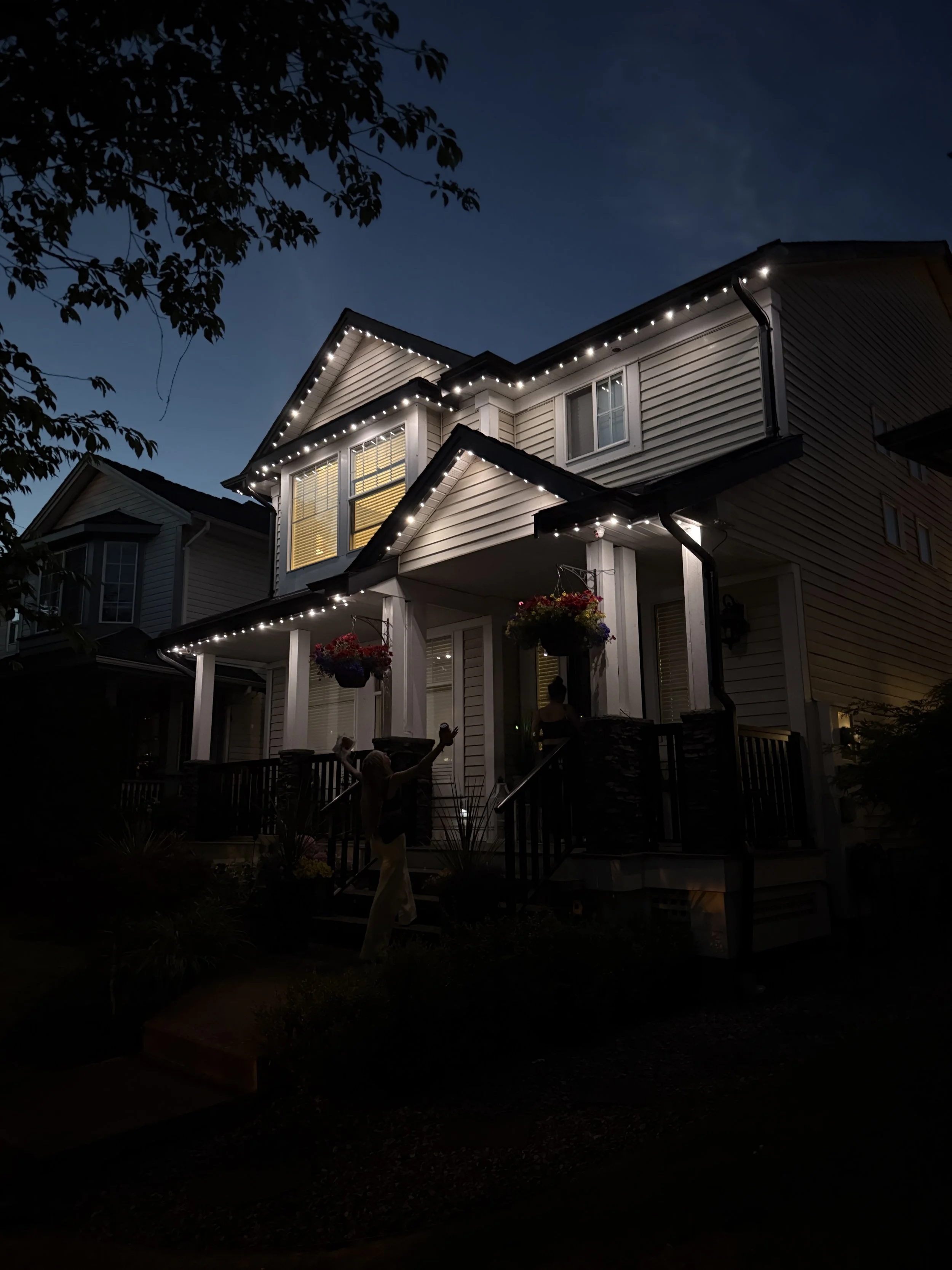 A two-story house decorated with string lights outlines on roof edges, front porch is lit with warm lighting, and there are hanging flower baskets, with people on and around the porch during dusk or night.
