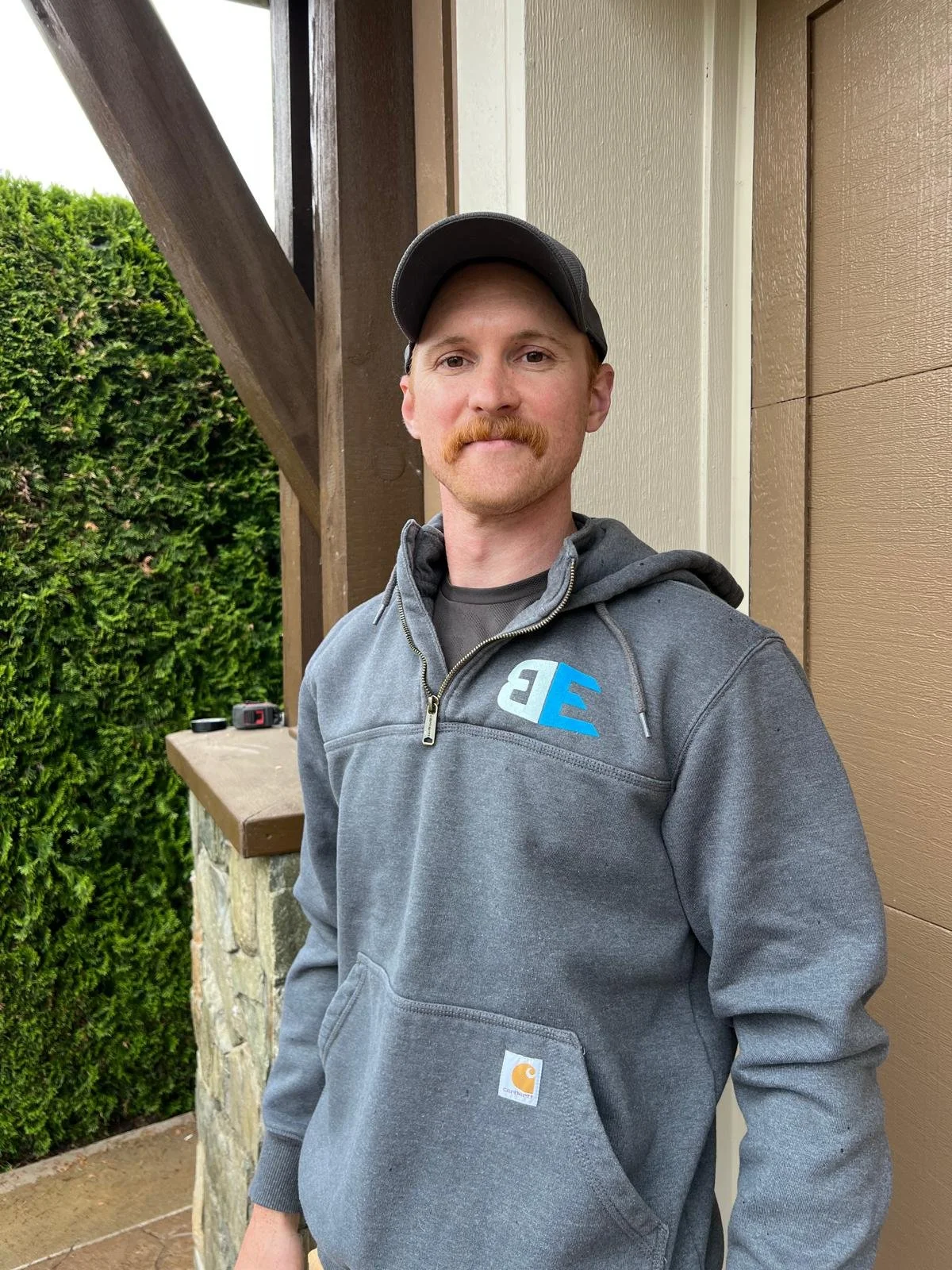 A man with a mustache and a baseball cap standing outside near a house, wearing a gray Carhartt hoodie with a logo on the chest, with greenery and a wooden structure in the background.