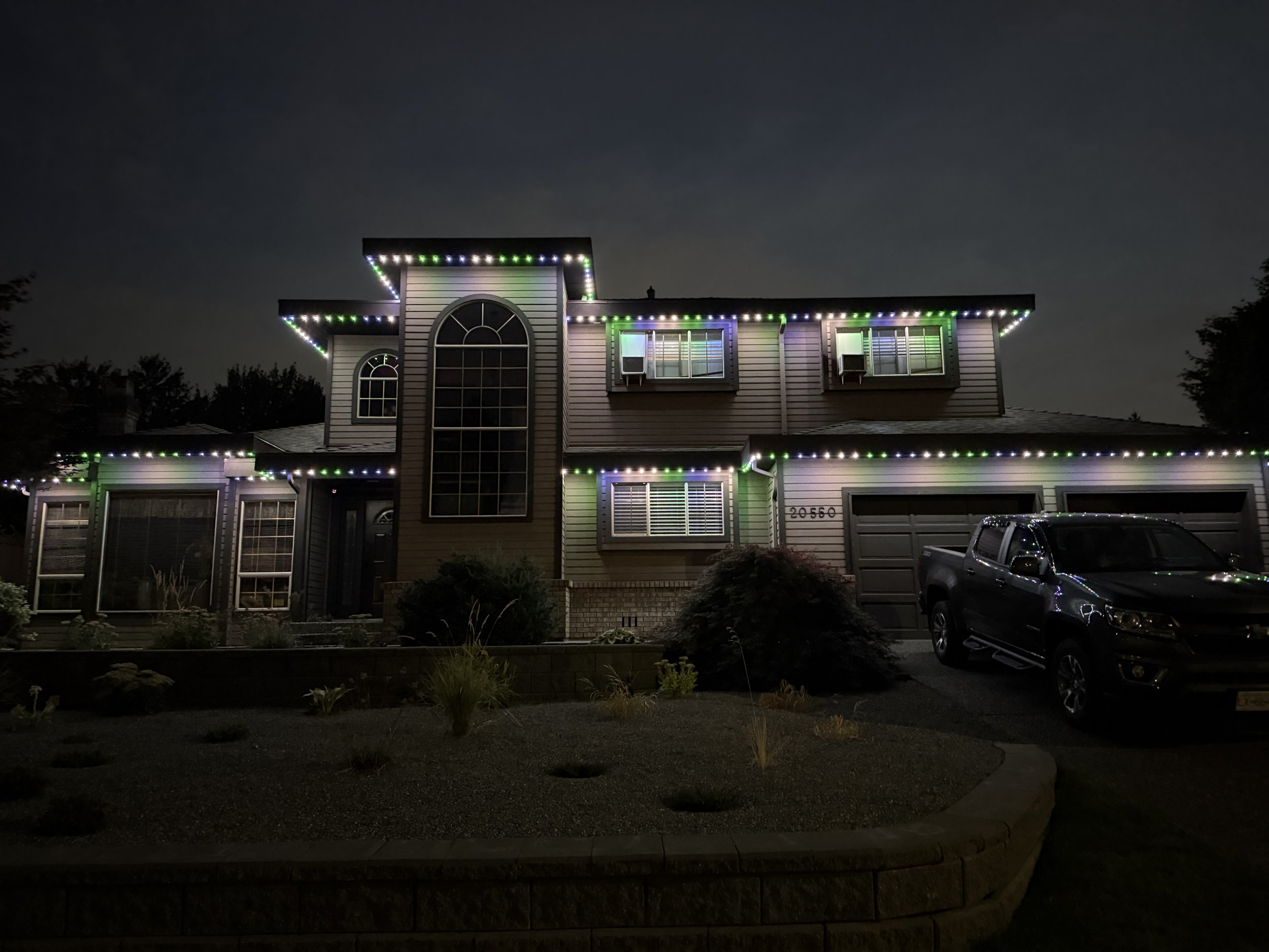 A two-story house decorated with colorful Christmas lights at night, with a black truck parked in the driveway.