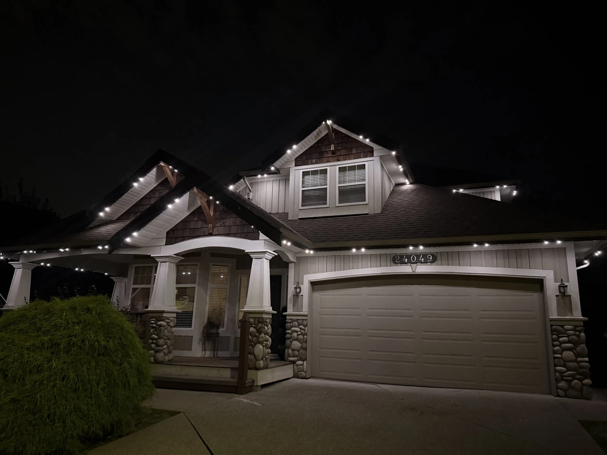 Nighttime photo of a house decorated with string lights on the roof and around the porch. The house has a garage, a small front porch, and a bush in front.