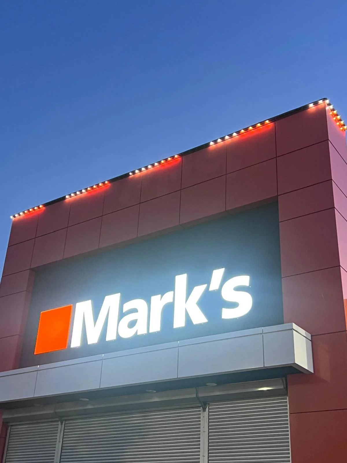 Close-up of a retail store's signage displaying the name 'Mark's' on a building with pink exterior wall and decorative lights at dusk.